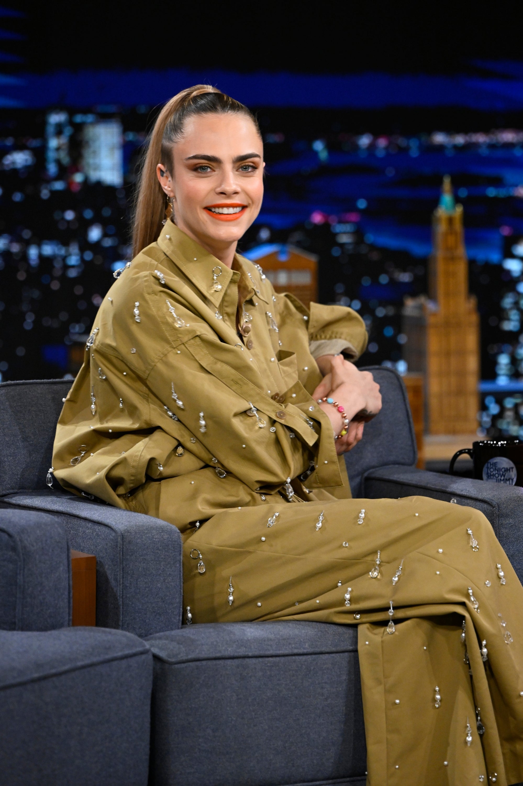 Cara Delevigne seated on a talk show set, wearing a loose-fitting outfit with embellishments