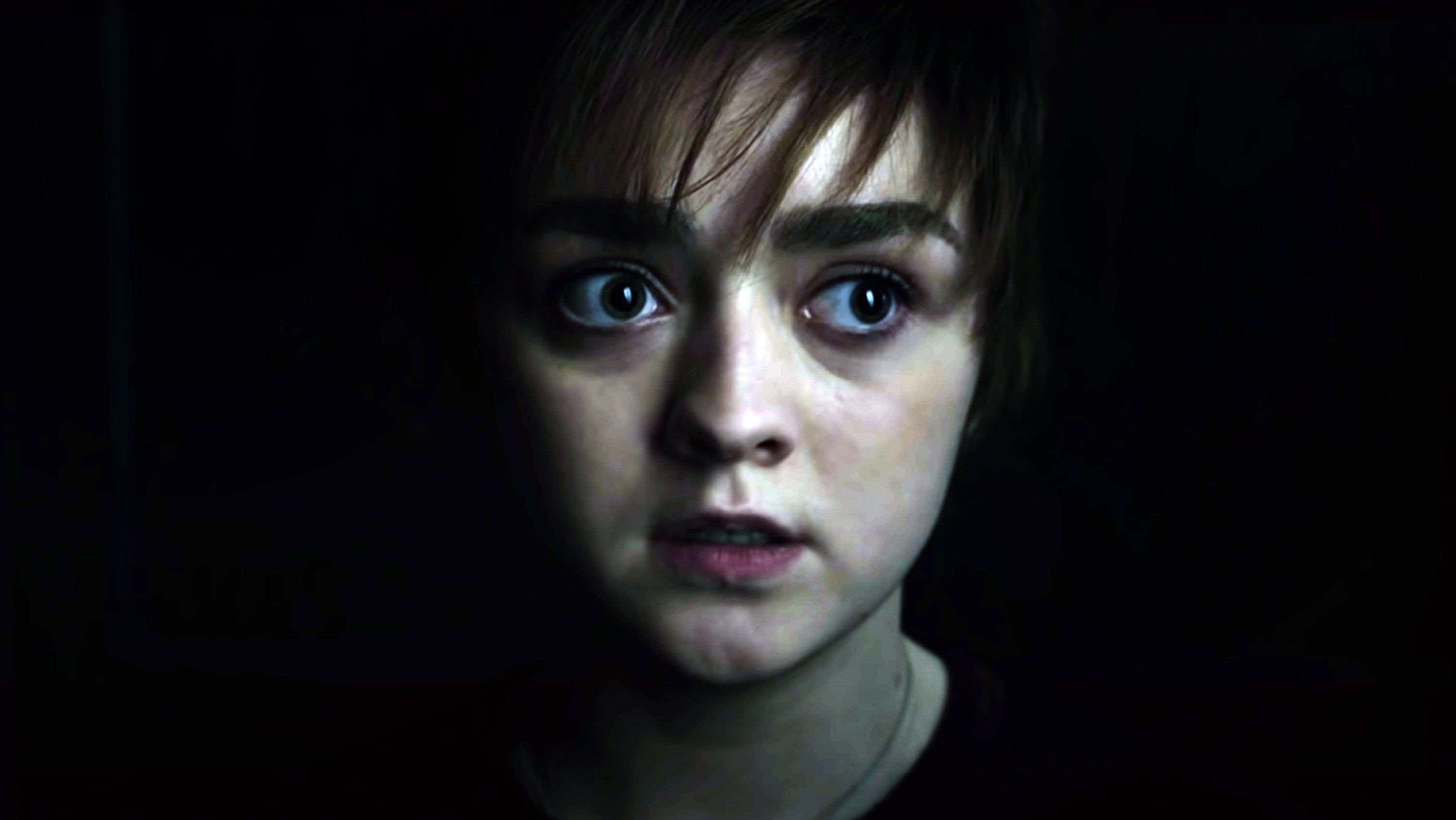 Maisie Williams with short hair, wide eyes, and a serious expression, looking slightly to the side in a dimly lit environment