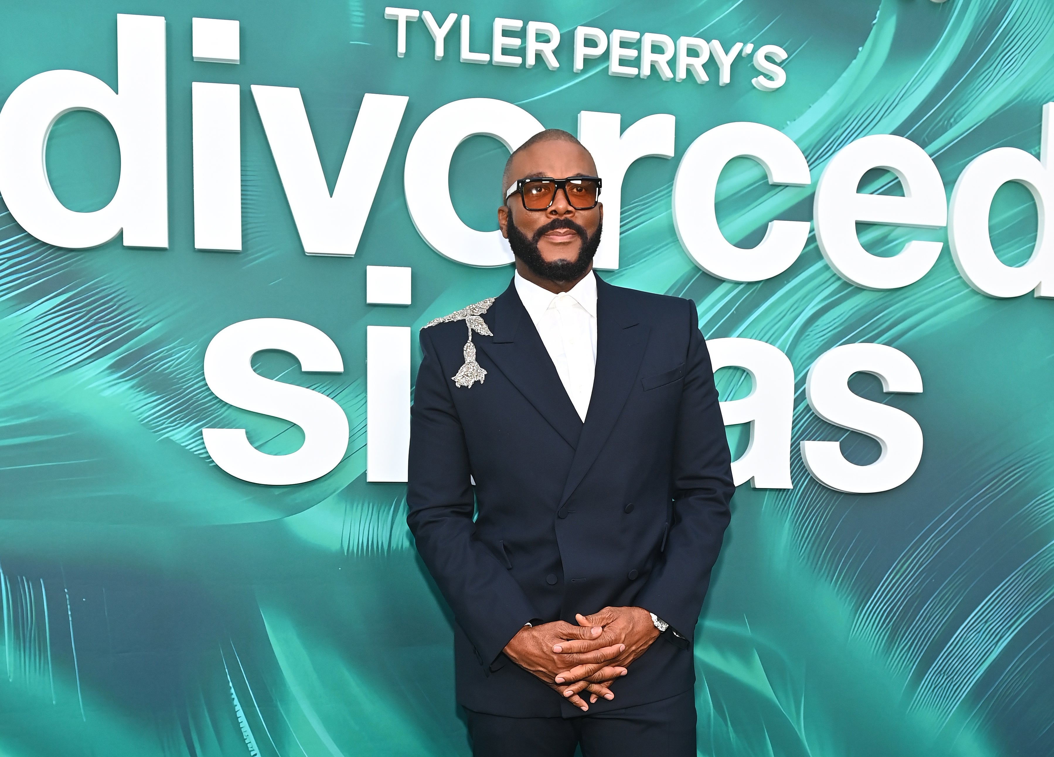 Tyler Perry in a tailored suit stands in front of a backdrop with "Tyler Perry's Divorced Silas" text, posing confidently