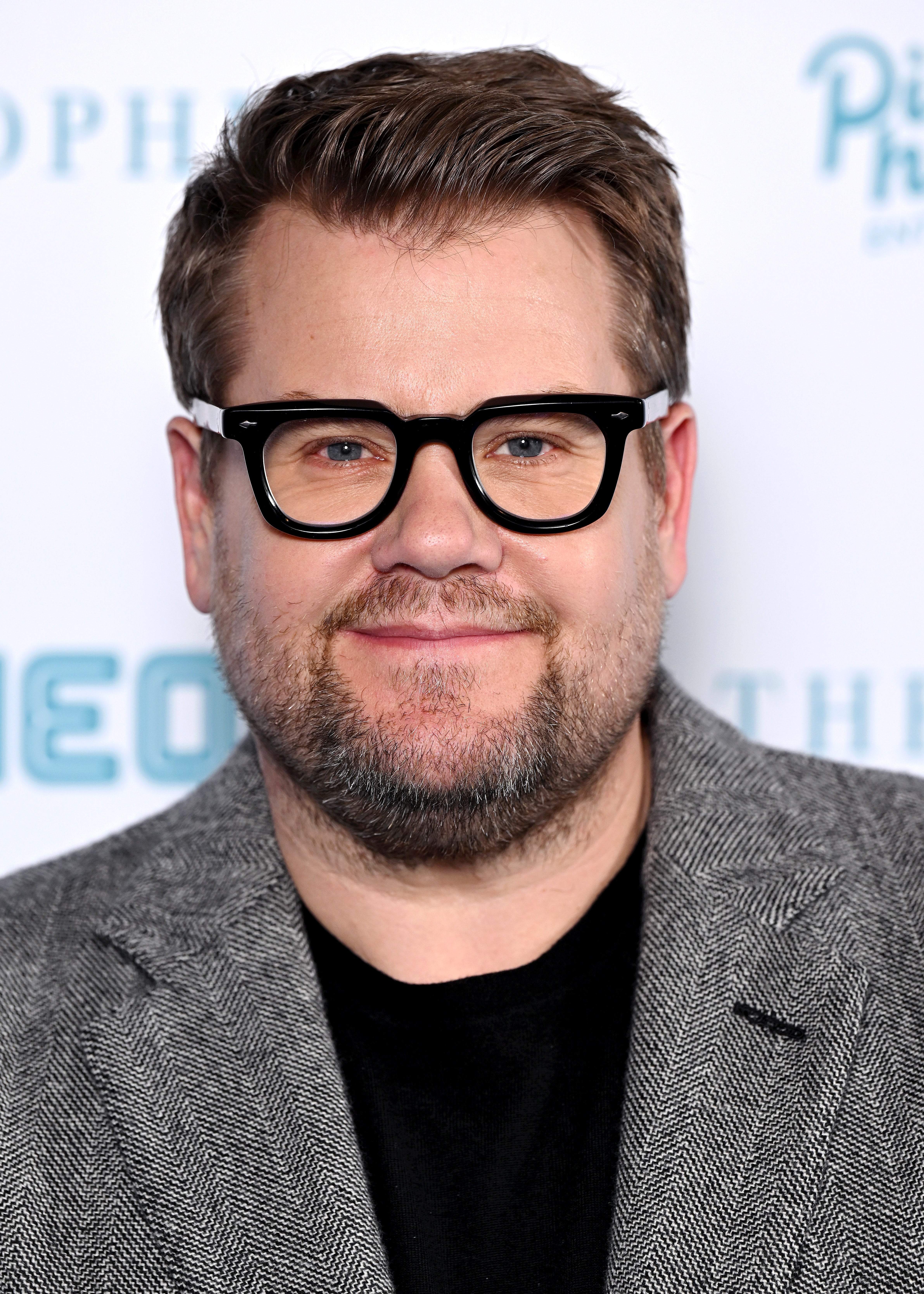 James Corden wearing glasses and a patterned blazer at a celebrity event, smiling at the camera
