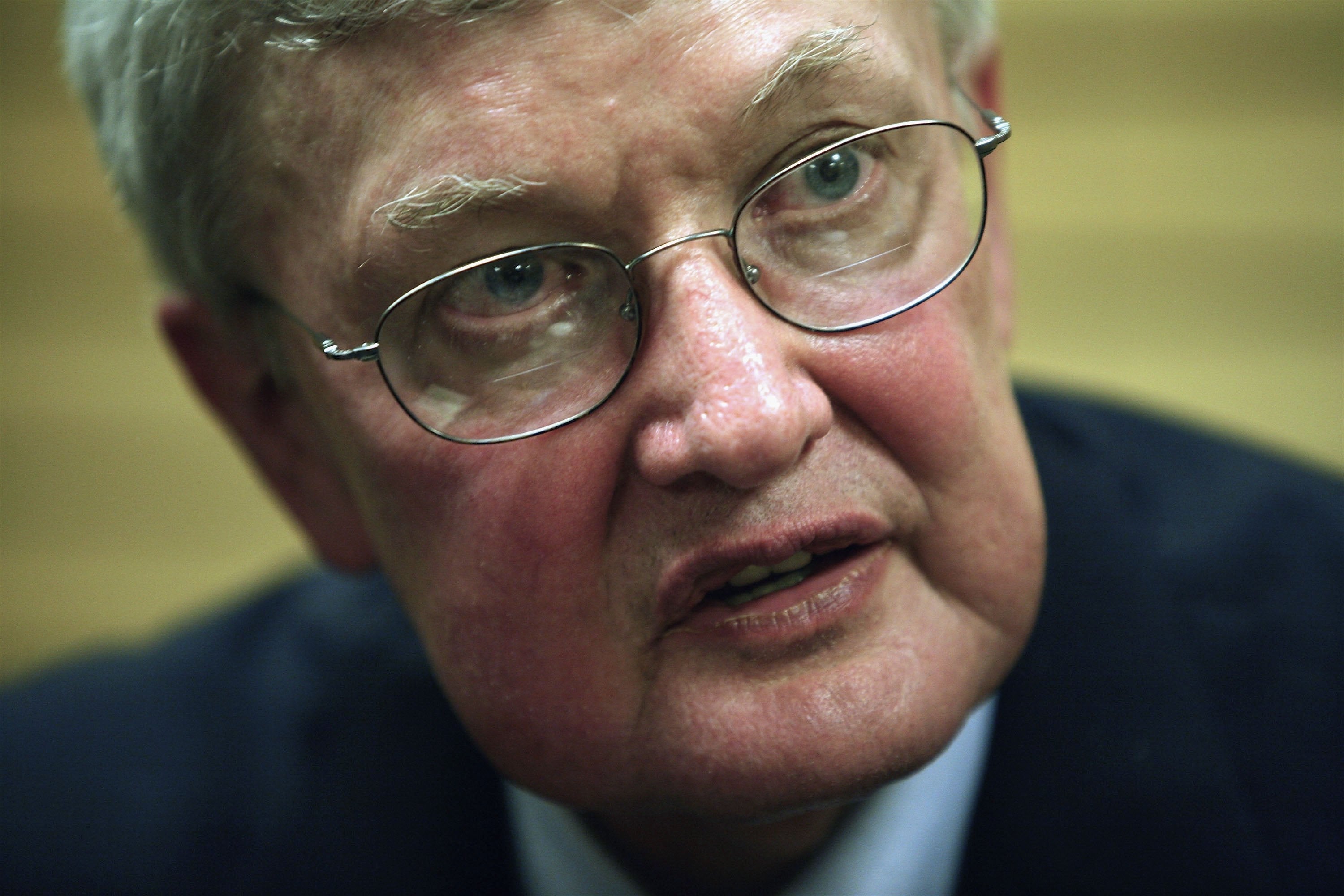 An older person with glasses and a thoughtful expression, wearing a dark suit jacket and light shirt