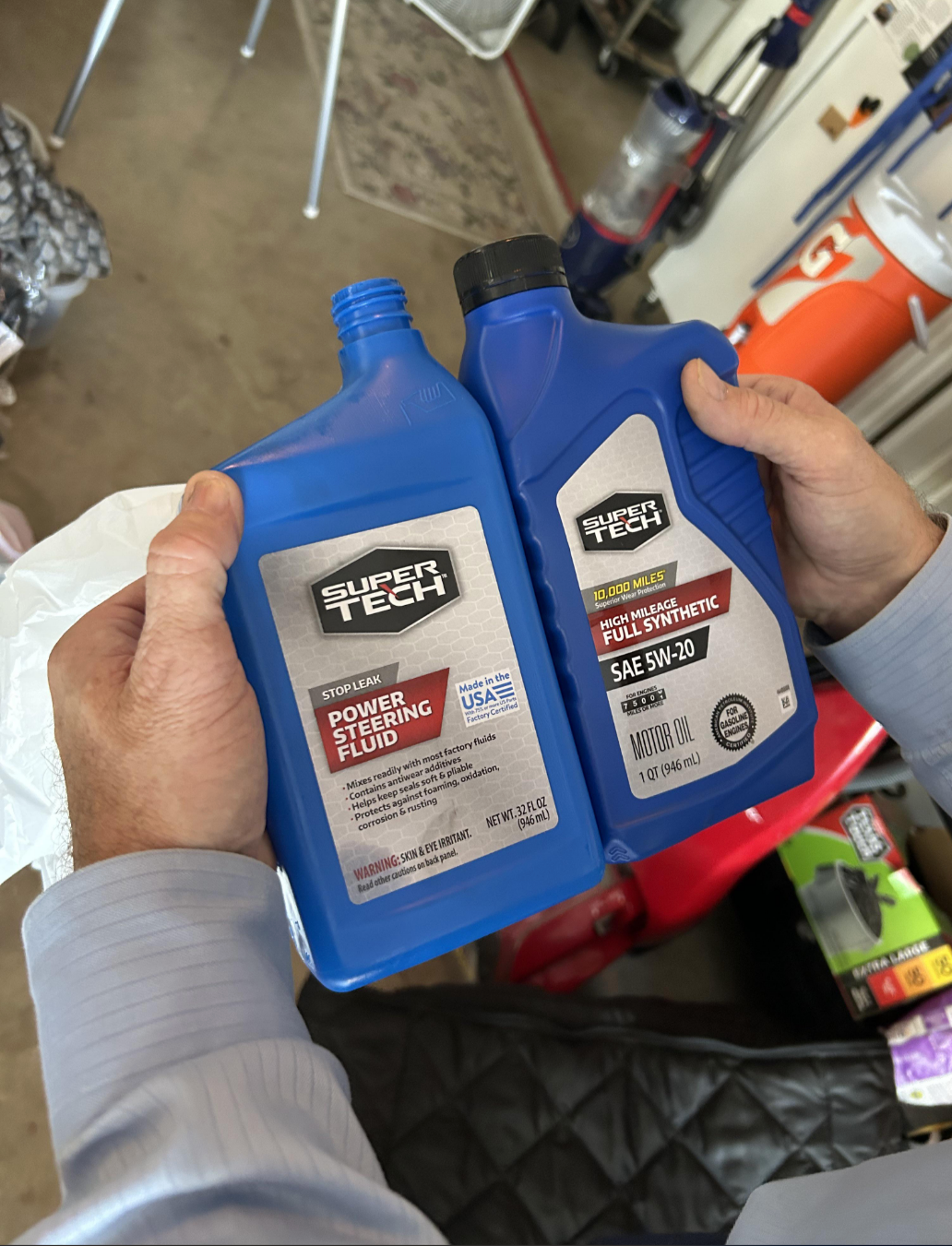Two hands holding blue bottles: one labeled "Power Steering Fluid" and the other "Full Synthetic Motor Oil SAE 5W-20"