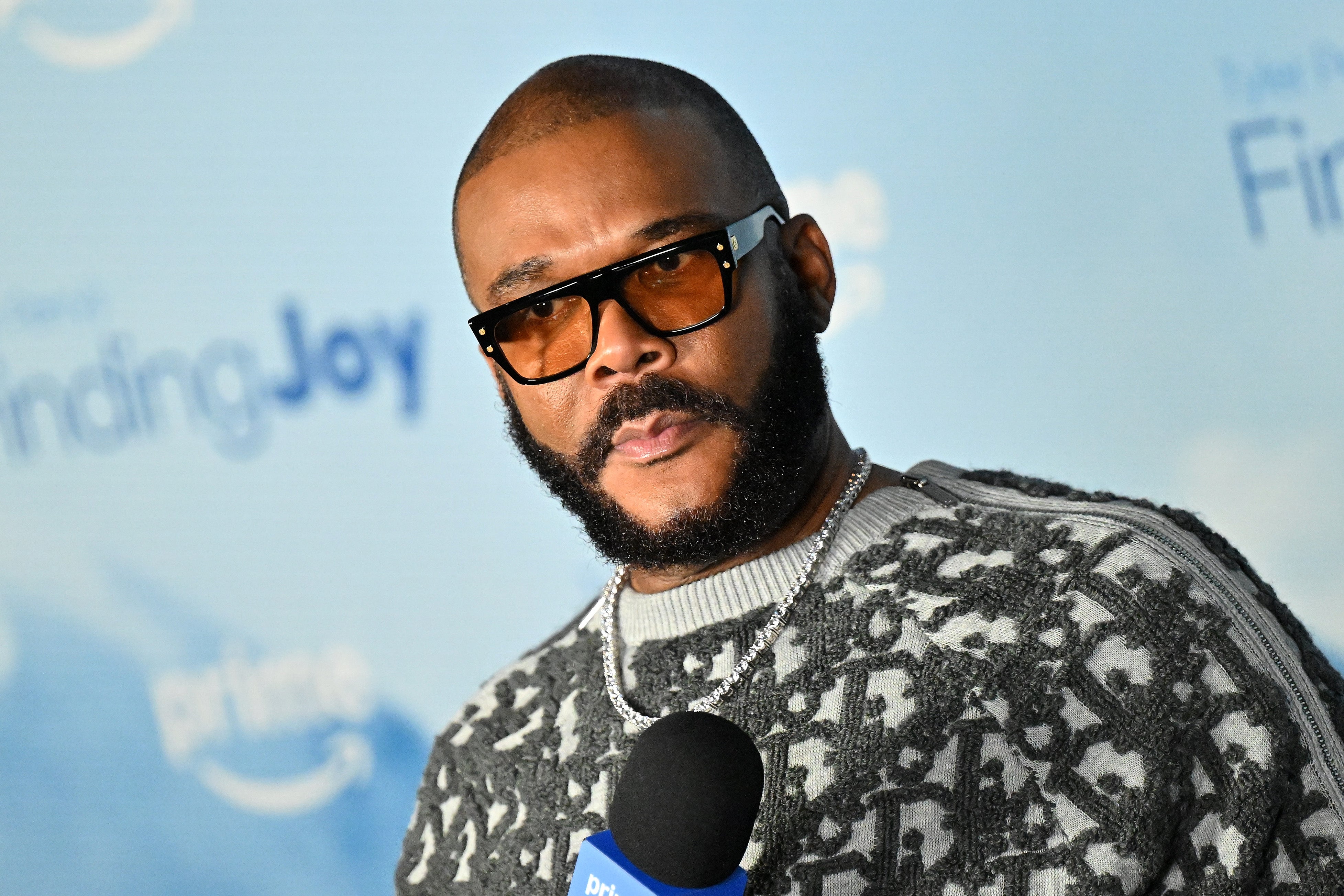 Tyler Perry wearing sunglasses and a patterned sweater speaks into a microphone at a media event