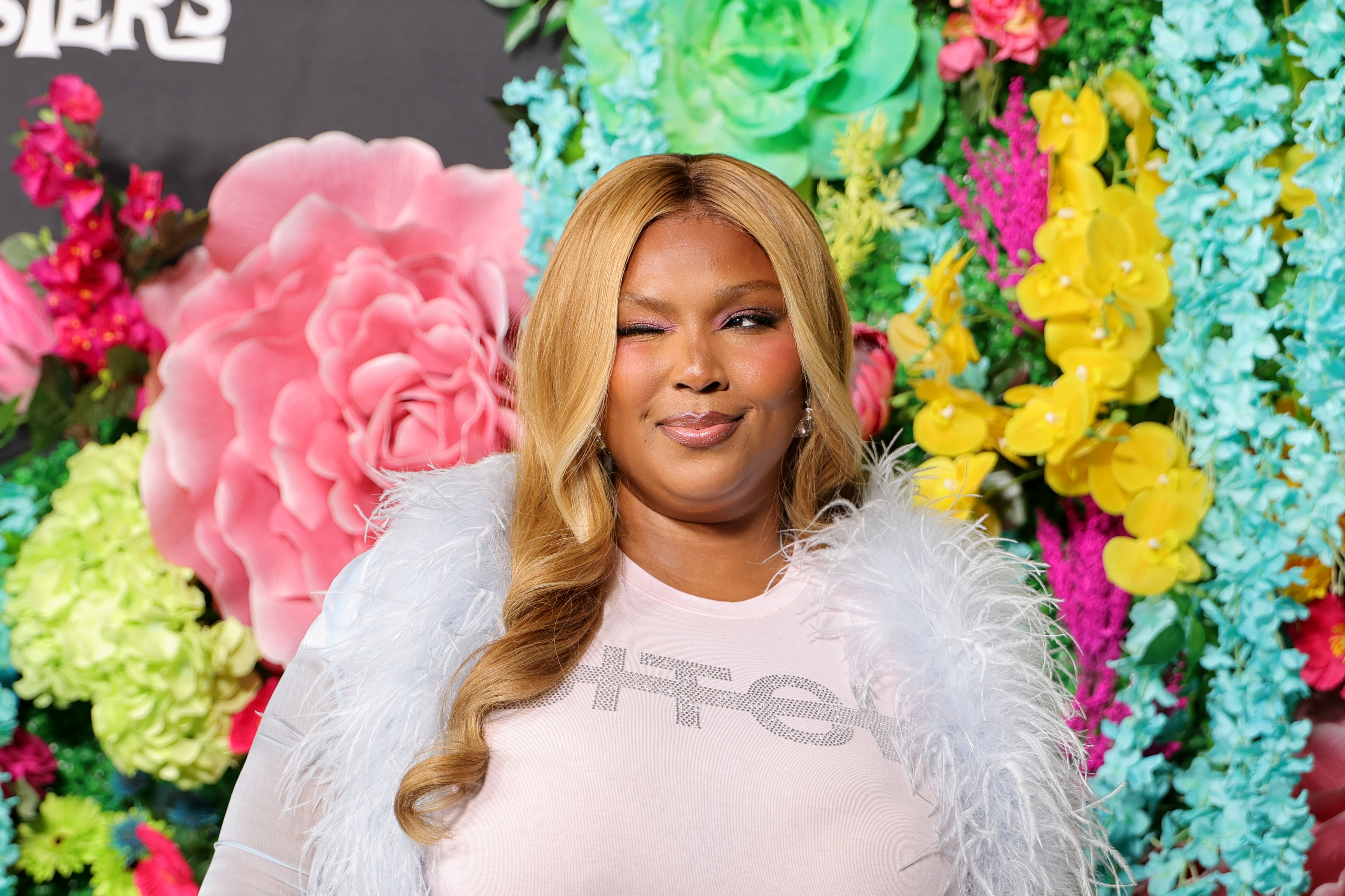 Lizzo winking at the camera, wearing a feathered outfit, in front of a vibrant floral background