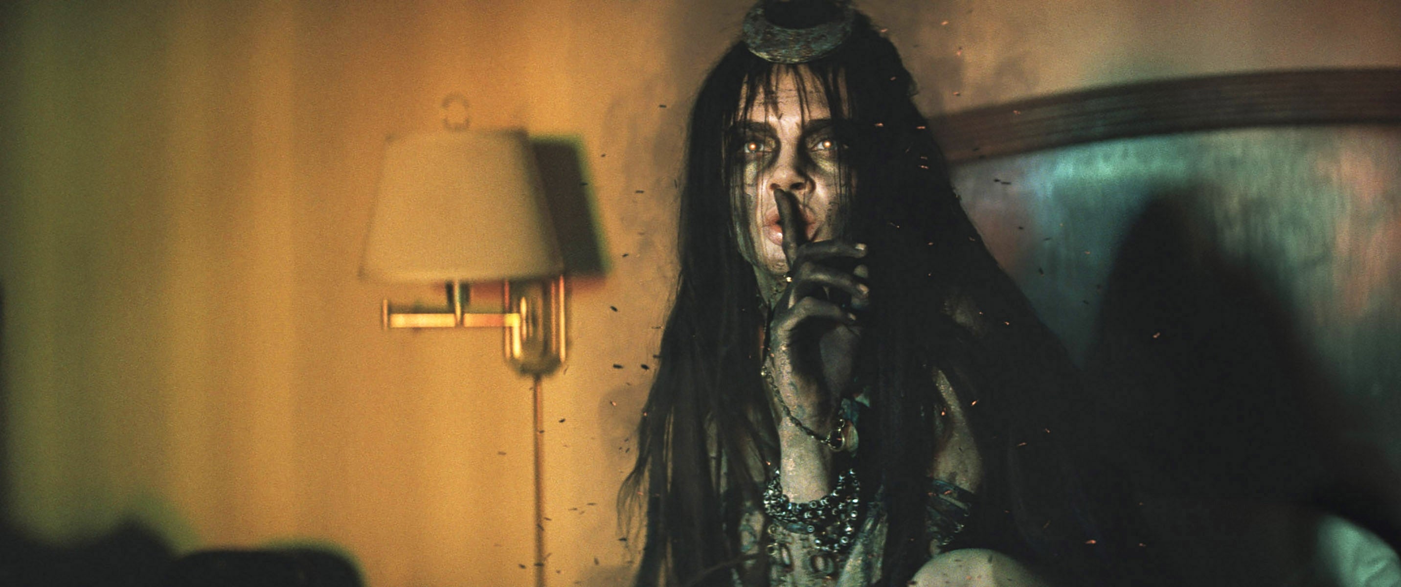 Cara Delevingne with long hair and tribal accessories holding a finger to their lips in a dimly lit setting in Suicide Squad
