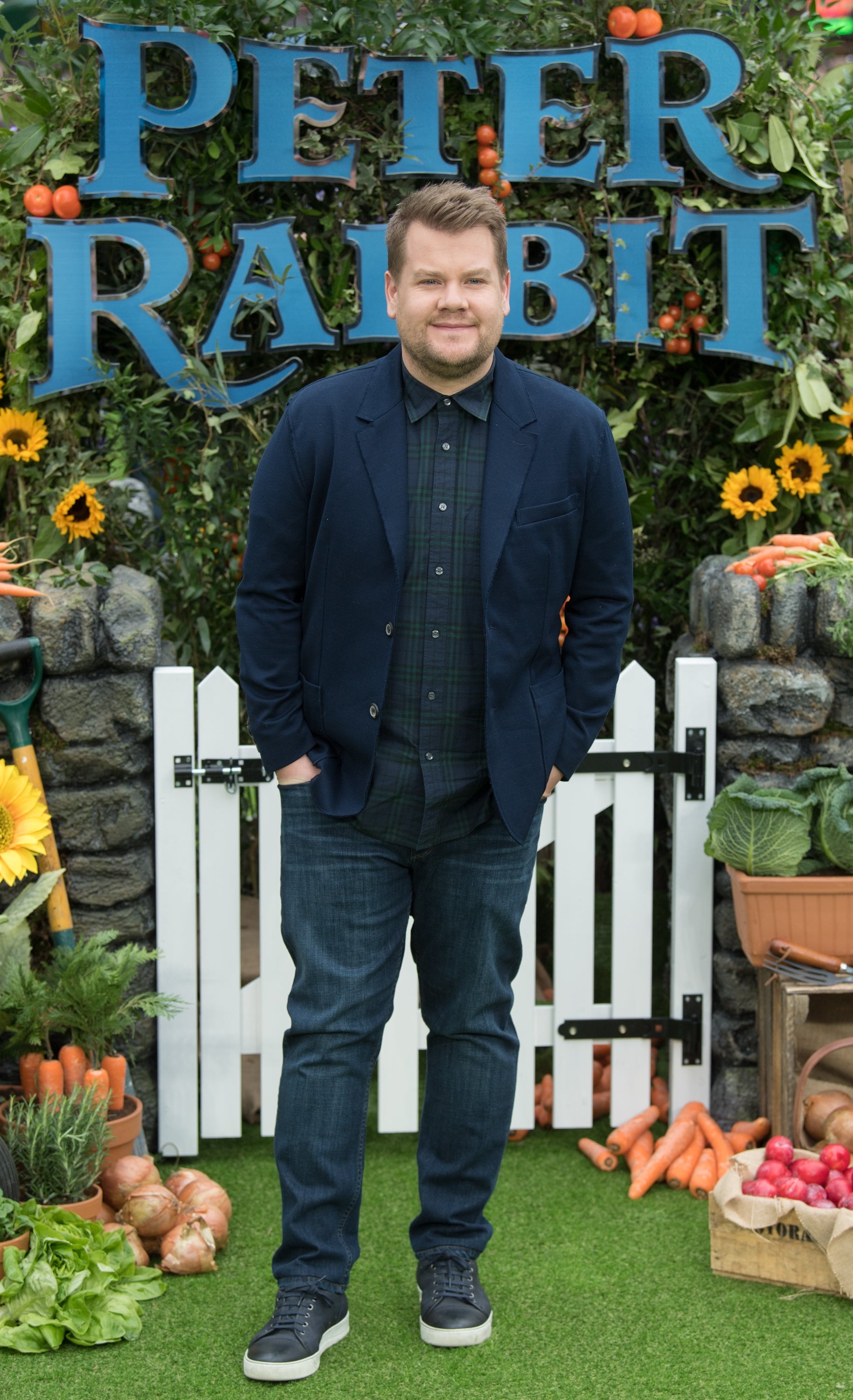 Person standing in front of "Peter Rabbit" sign, wearing a dark blazer, plaid shirt, and jeans, surrounded by garden-themed decor