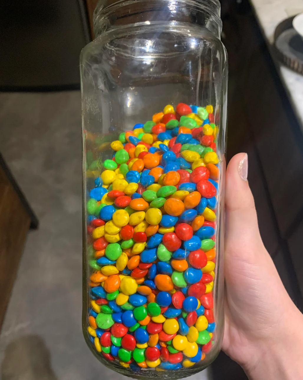 A jar filled with colorful small candies being held by a hand in a kitchen setting
