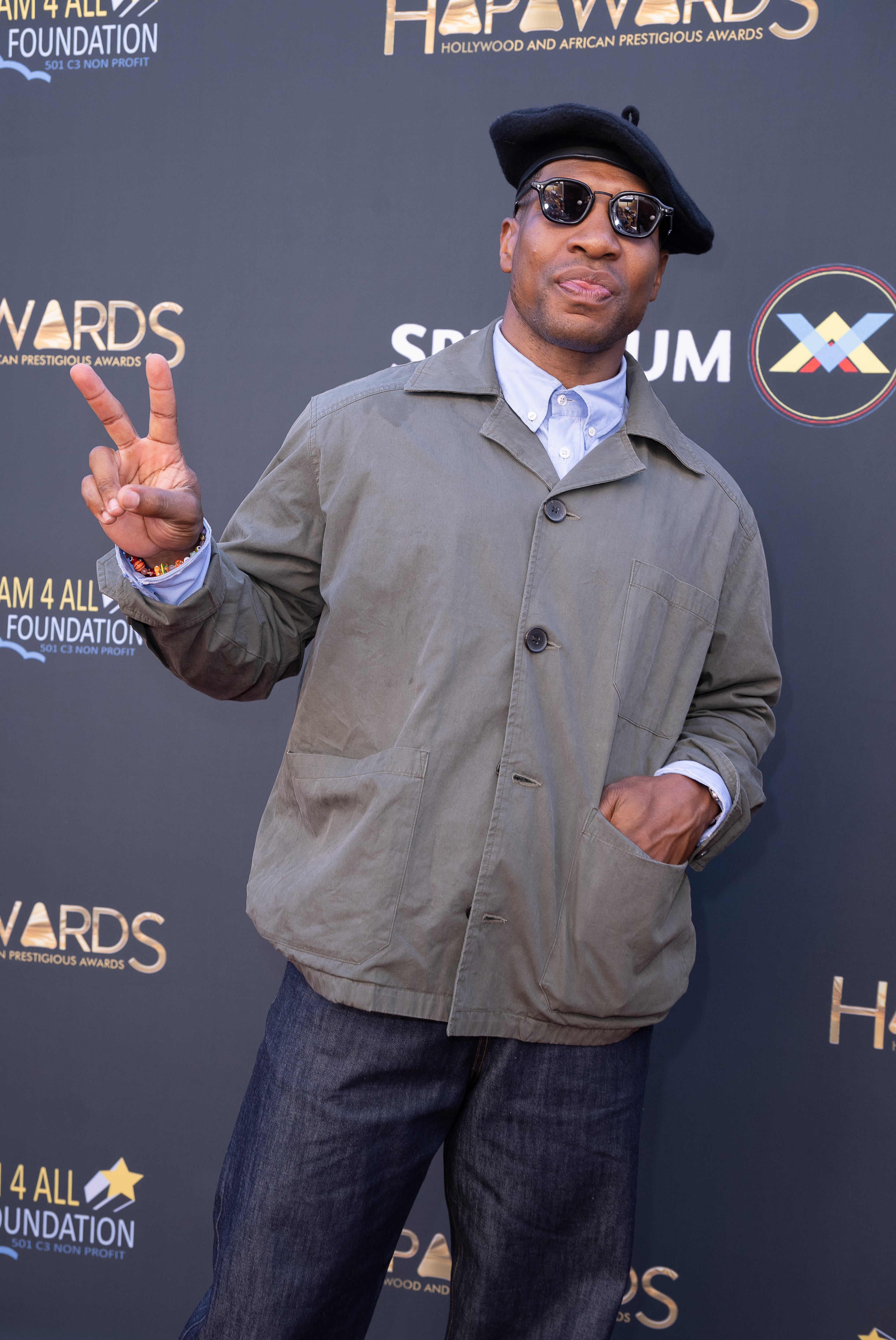 Jonathan Majors poses on a red carpet, wearing a casual jacket, button-up shirt, dark pants, sunglasses, and a hat, making a peace sign gesture