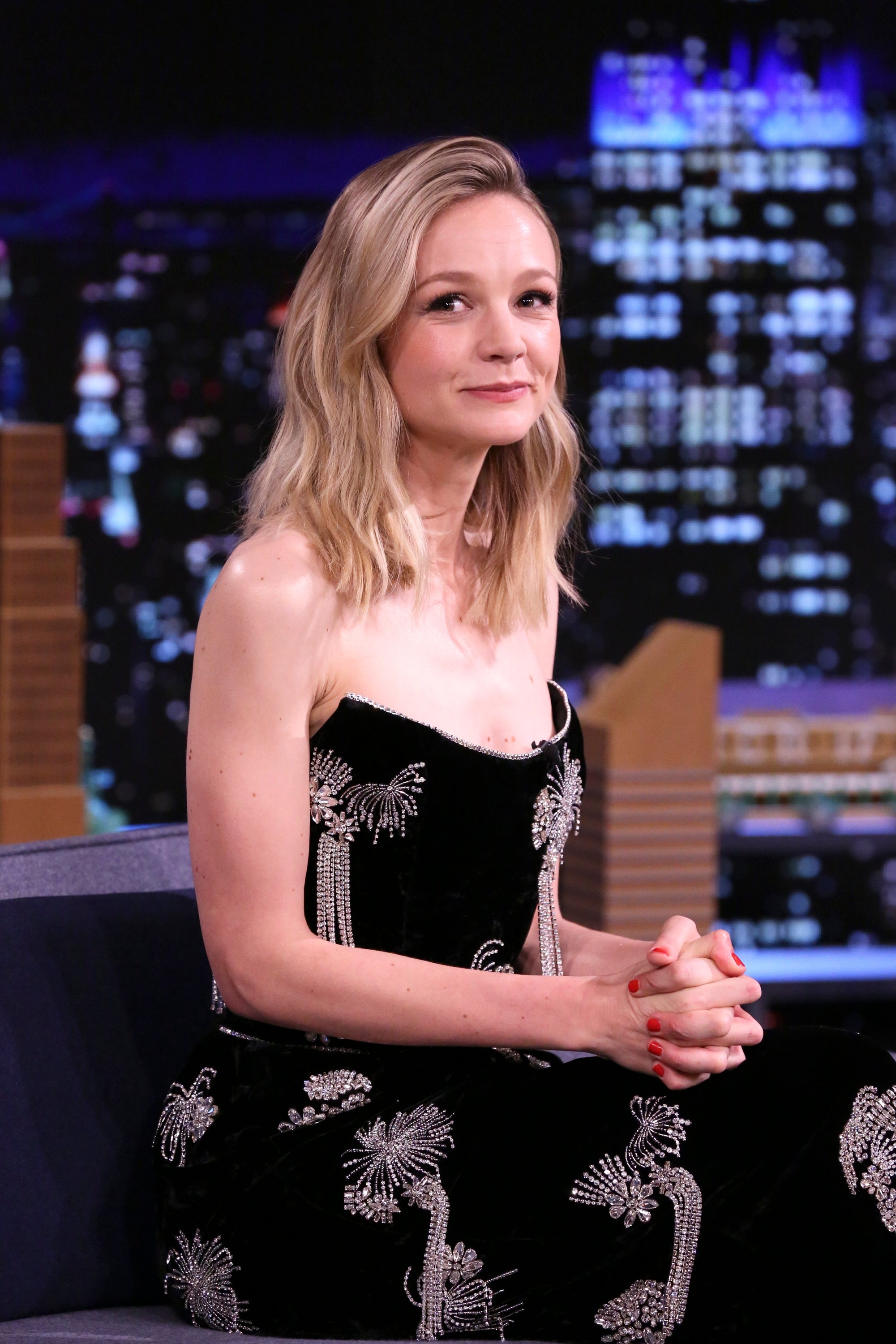 Carey Mulligan in an elegant, embellished gown sits on a talk show set, hands clasped