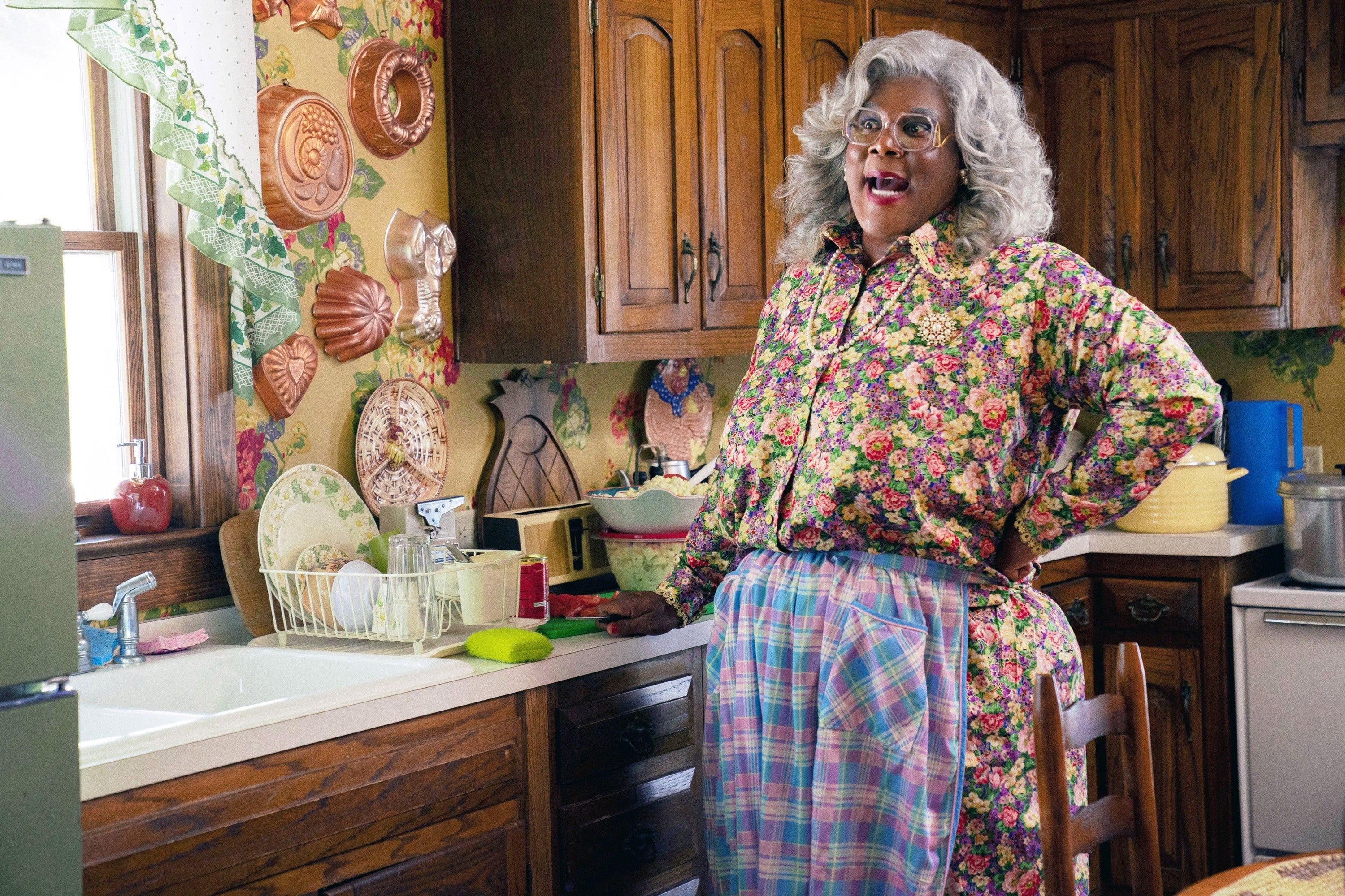Tyler Perry as Madea in a floral blouse and plaid skirt stands confidently in a vintage-style kitchen, exuding a humorous and lively presence