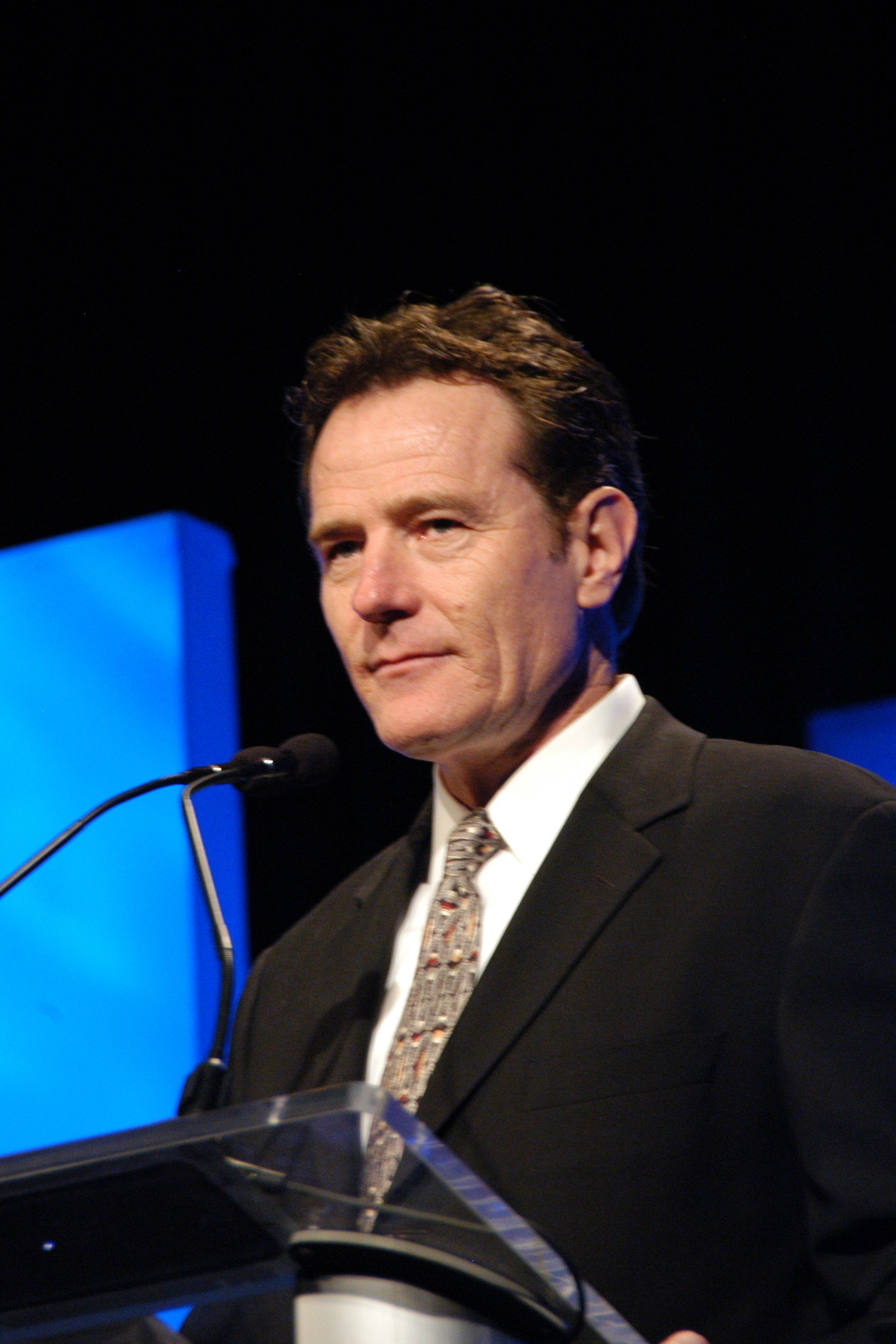 Bryan Cranston in a suit and tie speaks at a podium on stage