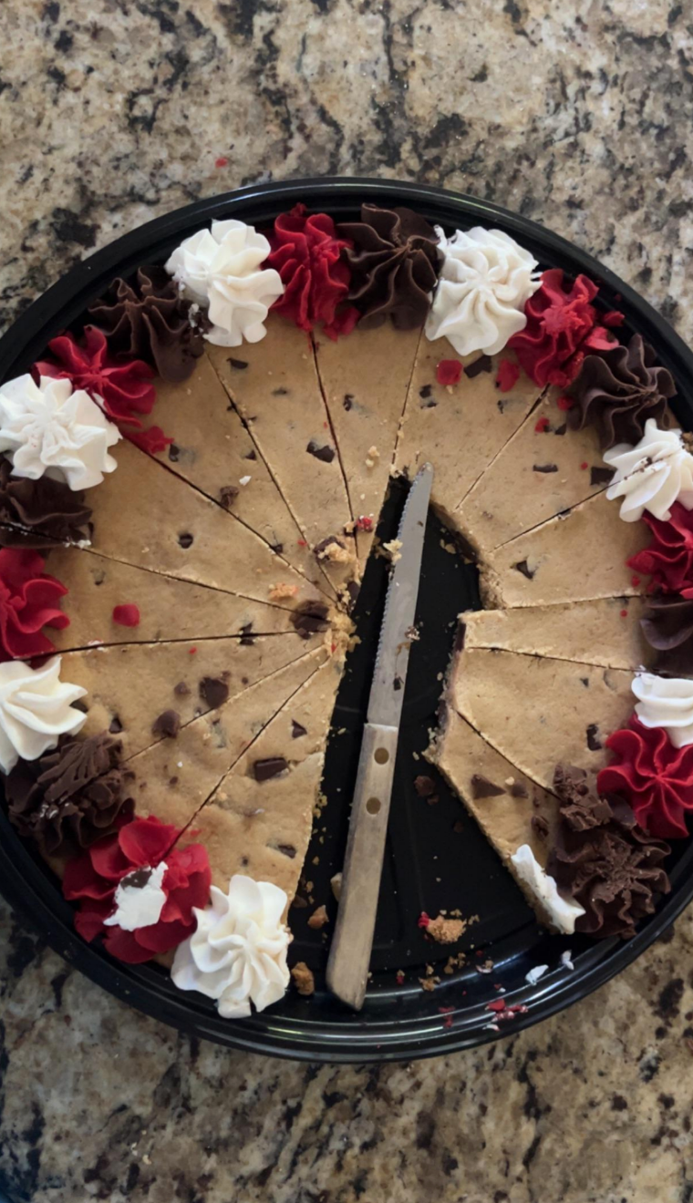 A large cookie cake with slices missing, topped with swirls of white, red, and chocolate frosting, and a knife placed on the tray