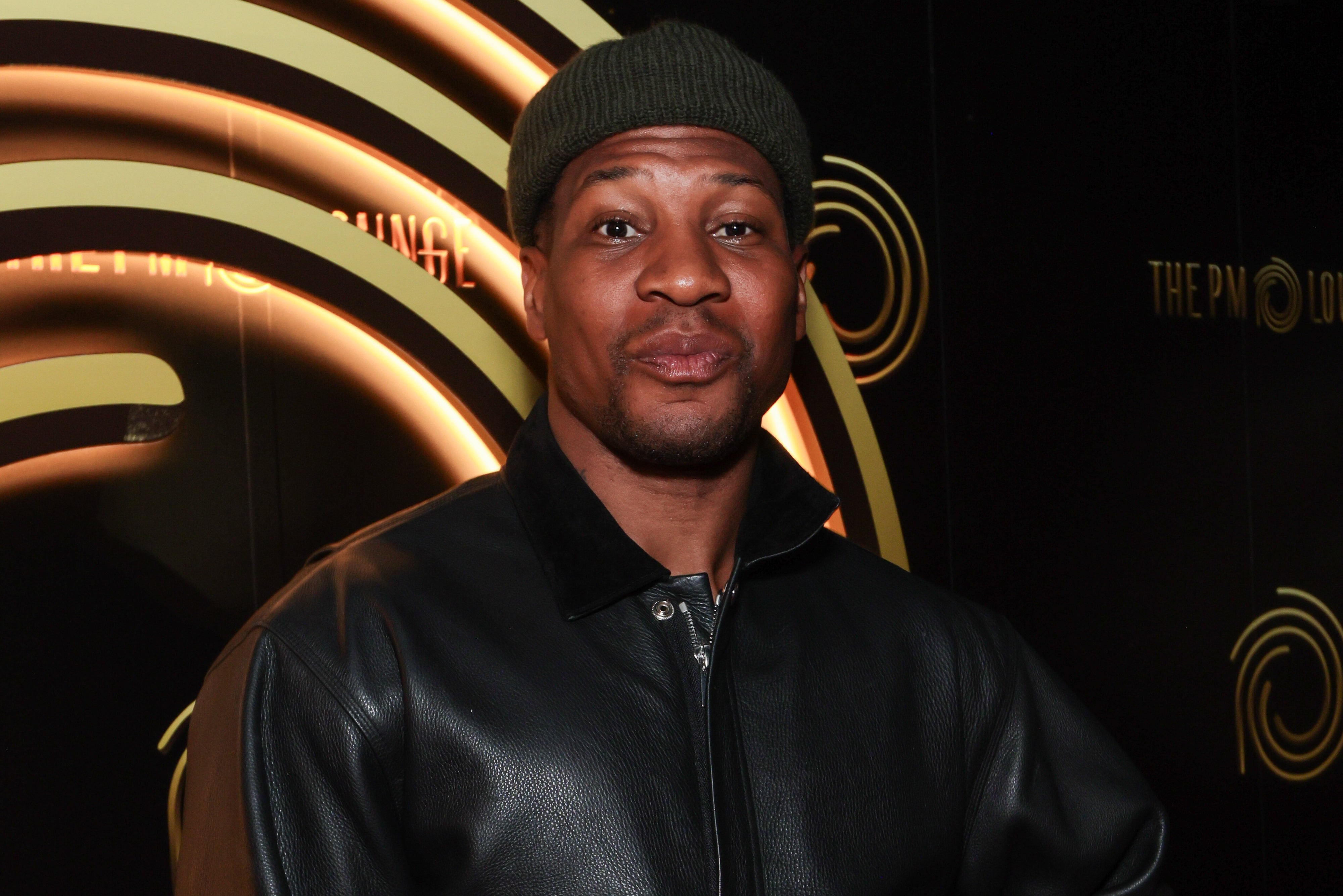 Jonathan Majors in a beanie and leather jacket poses confidently against a backdrop with circular designs at an event