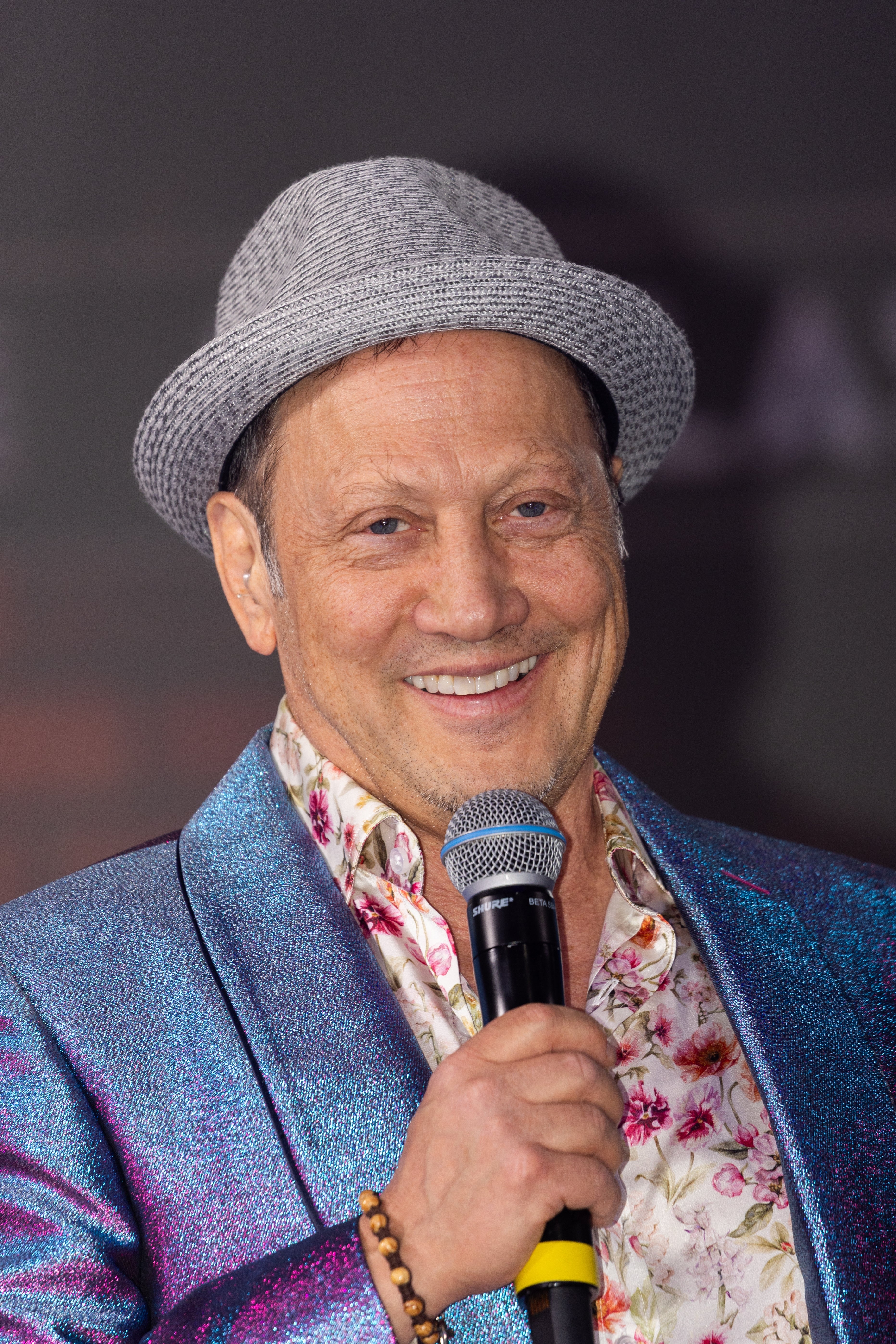 Rob Schneider wearing a shiny suit jacket, floral shirt, and a light textured hat, holding a microphone and smiling at an event