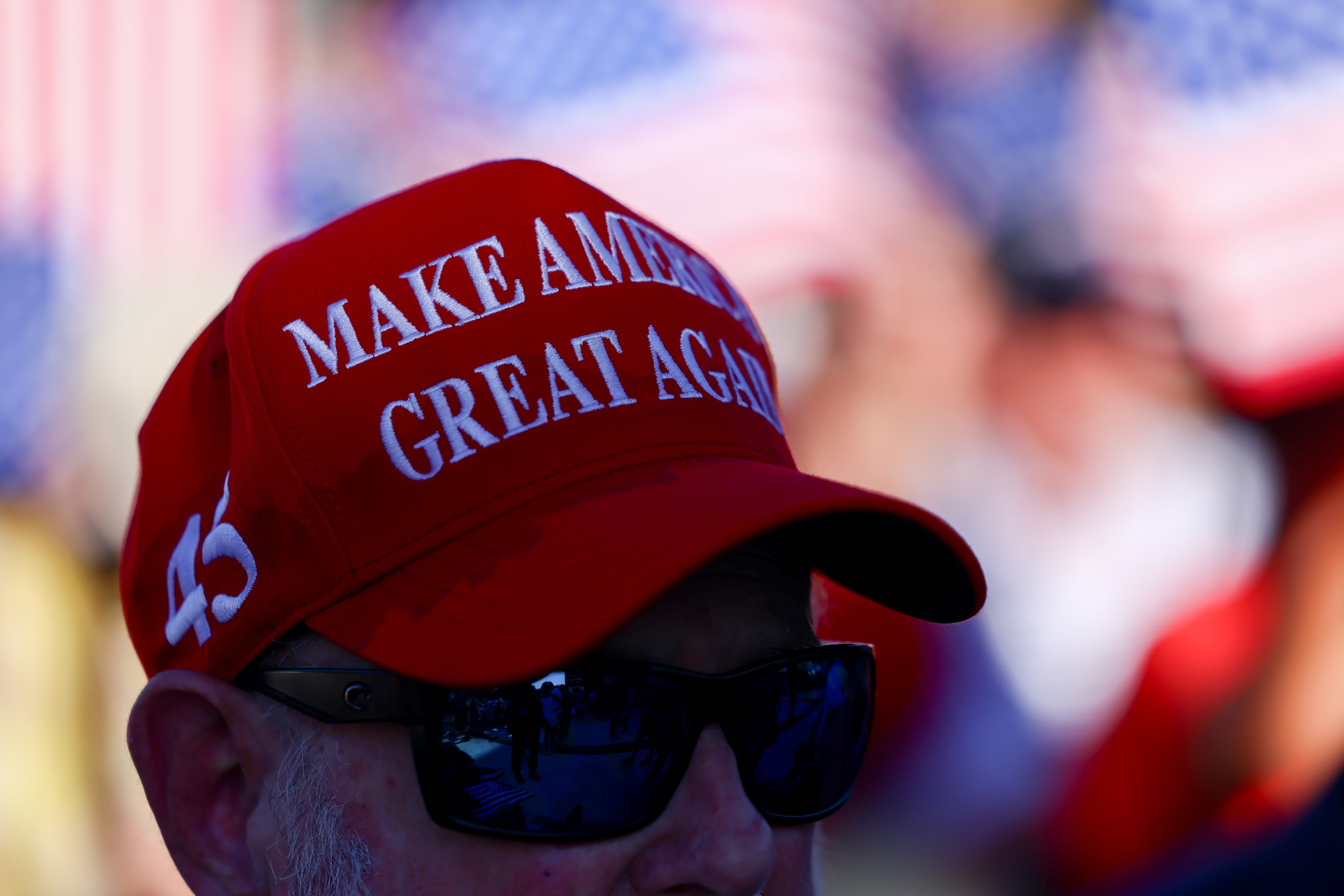Person wearing a "Make America Great Again" headdress with sunglasses, American flags successful the background