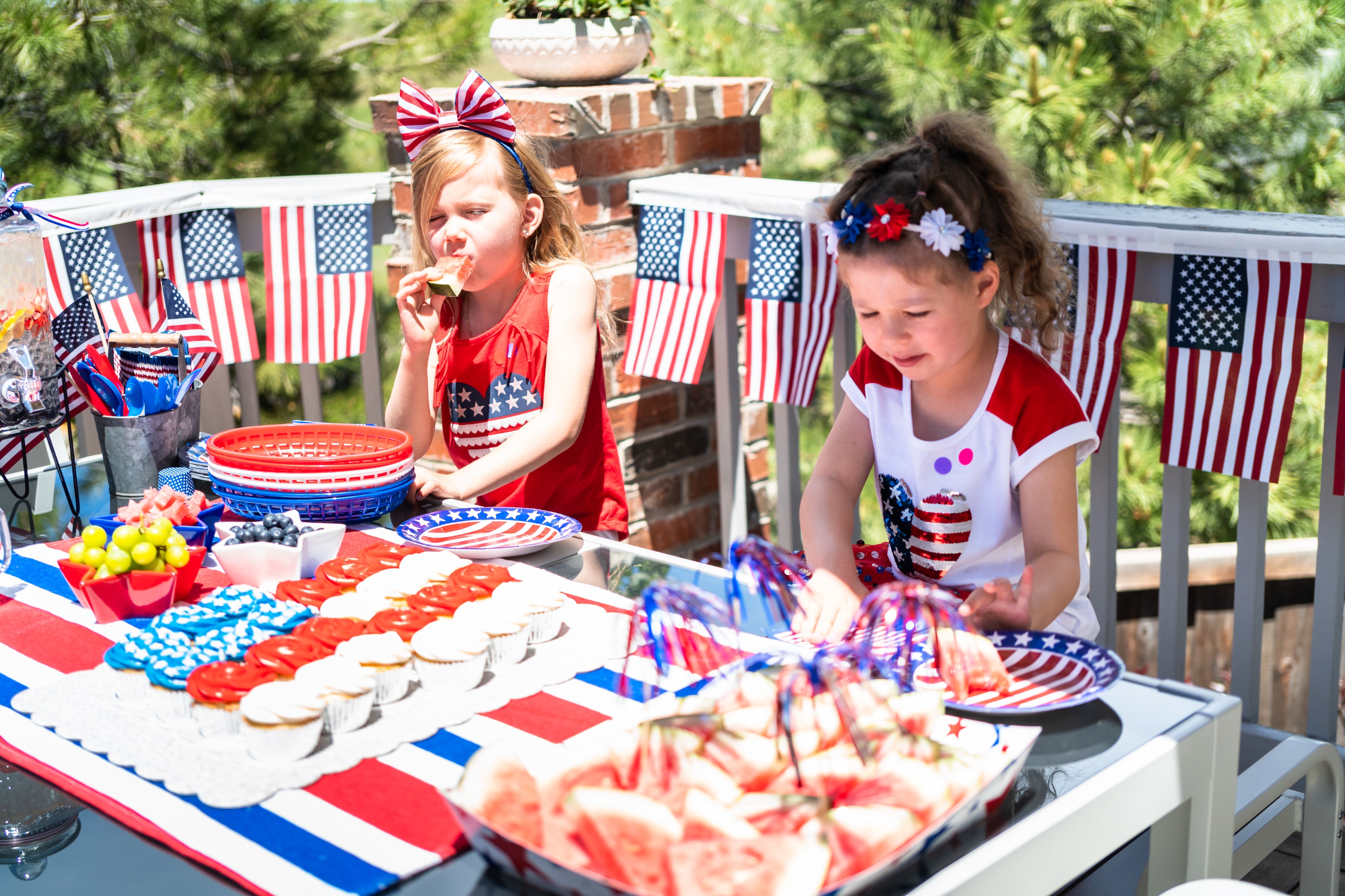 Two kids with patriotic outfits bask a array filled with festive red, white, and bluish themed nutrient outdoors, with American flags successful the background