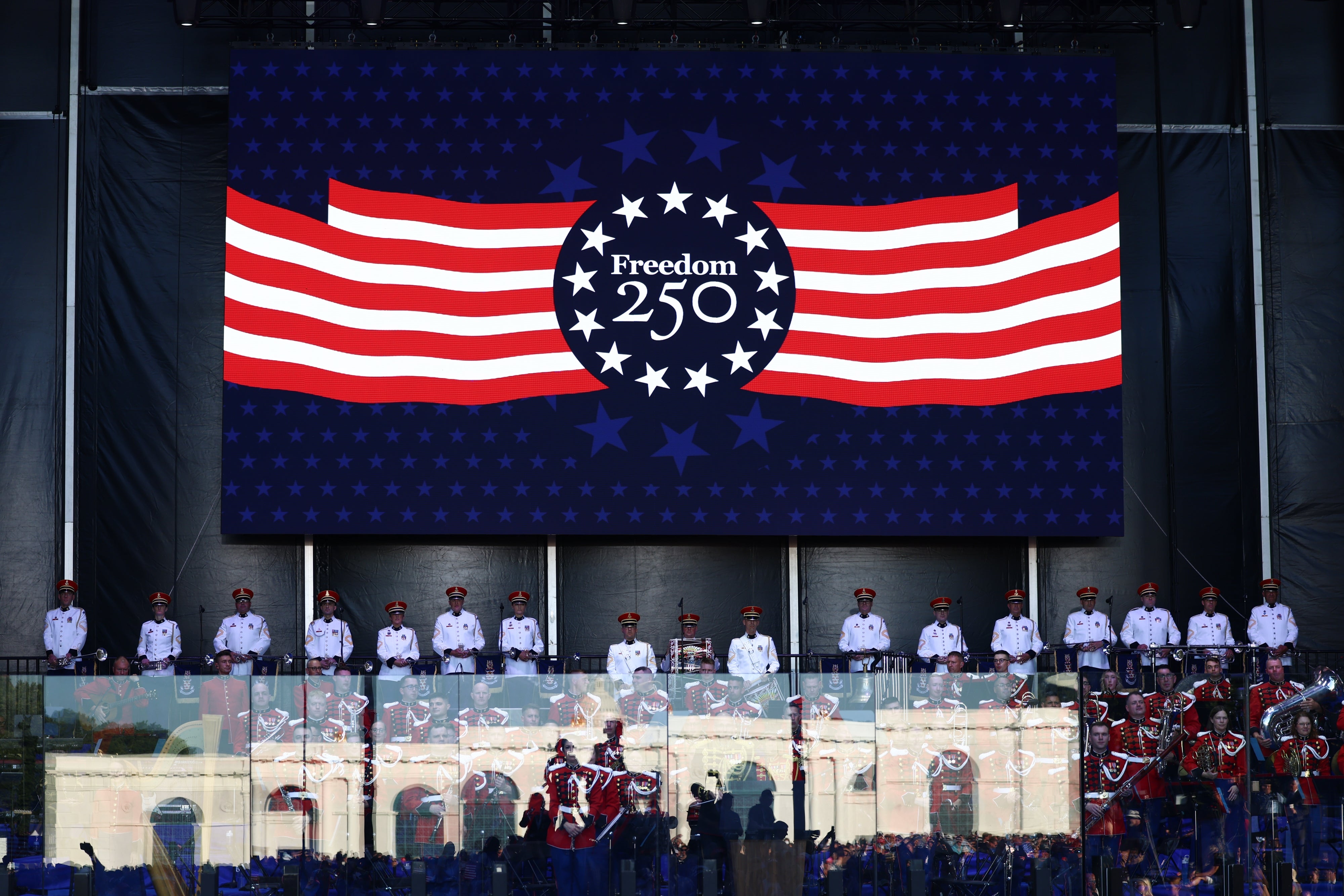 A banner reads "Freedom 250" with stars and stripes connected stage, supra uniformed individuals and musicians performing below