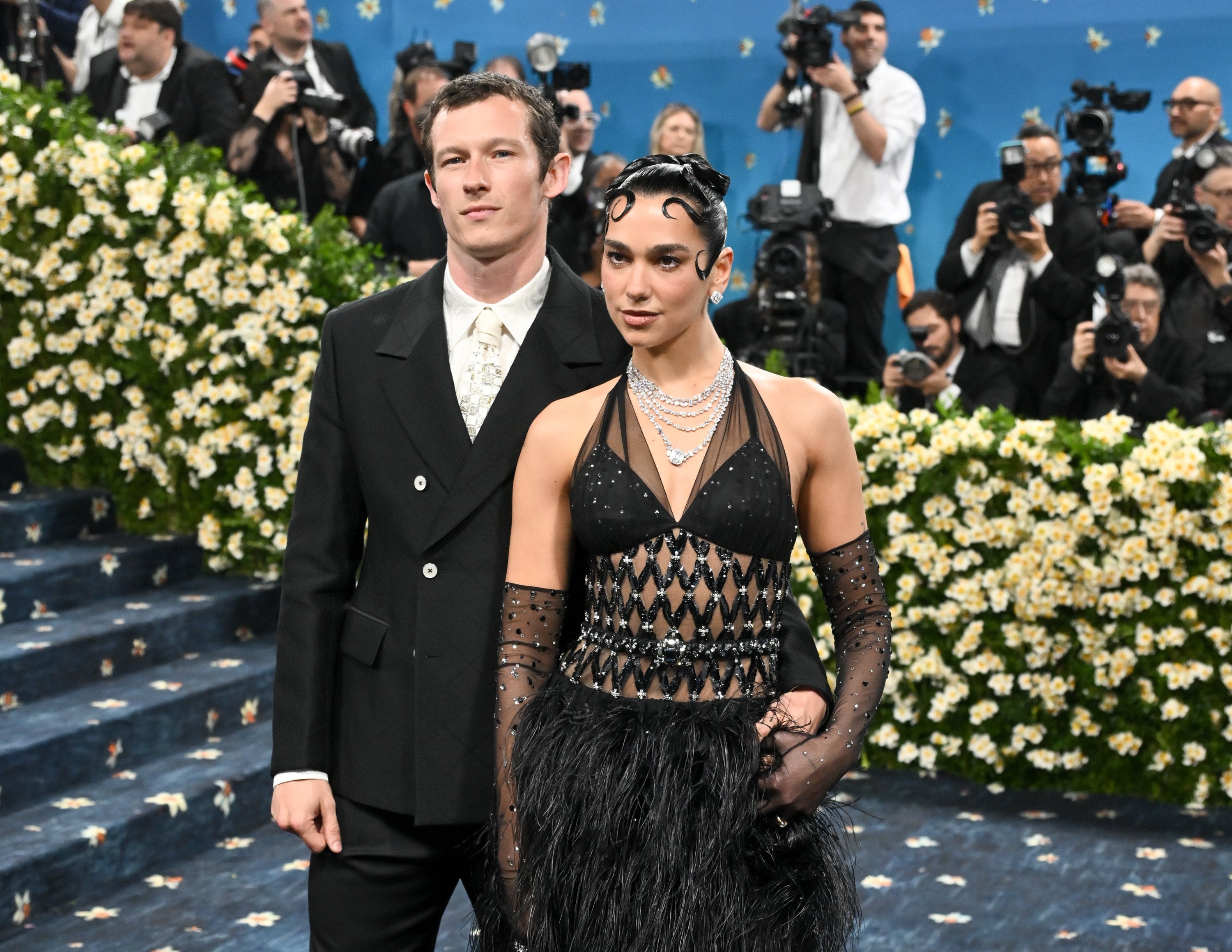 Two individuals pose together at a formal event. The woman wears a patterned gown with a feathered skirt, and the man is in a classic black suit