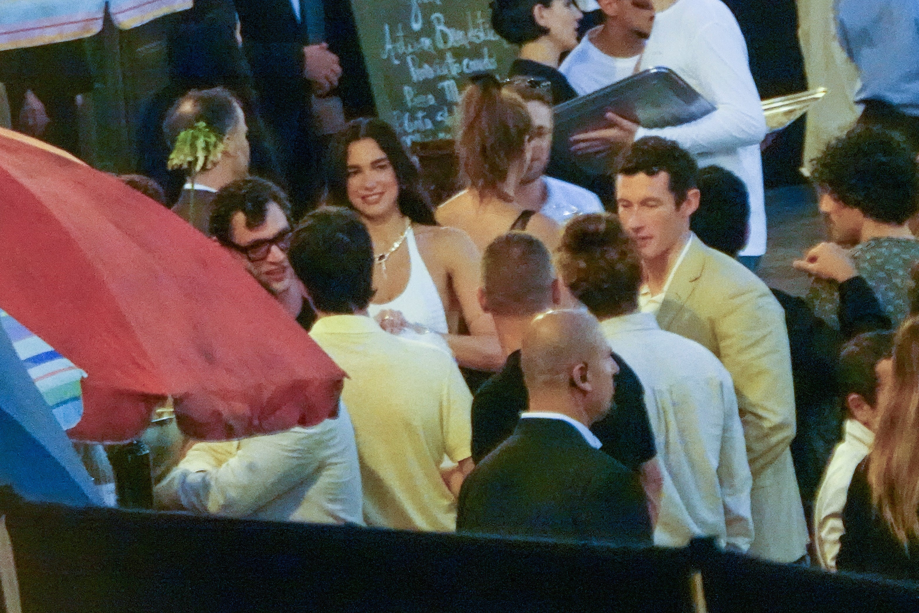 People socializing outdoors at a busy evening event; one individual in a white halter top stands out amidst the crowd