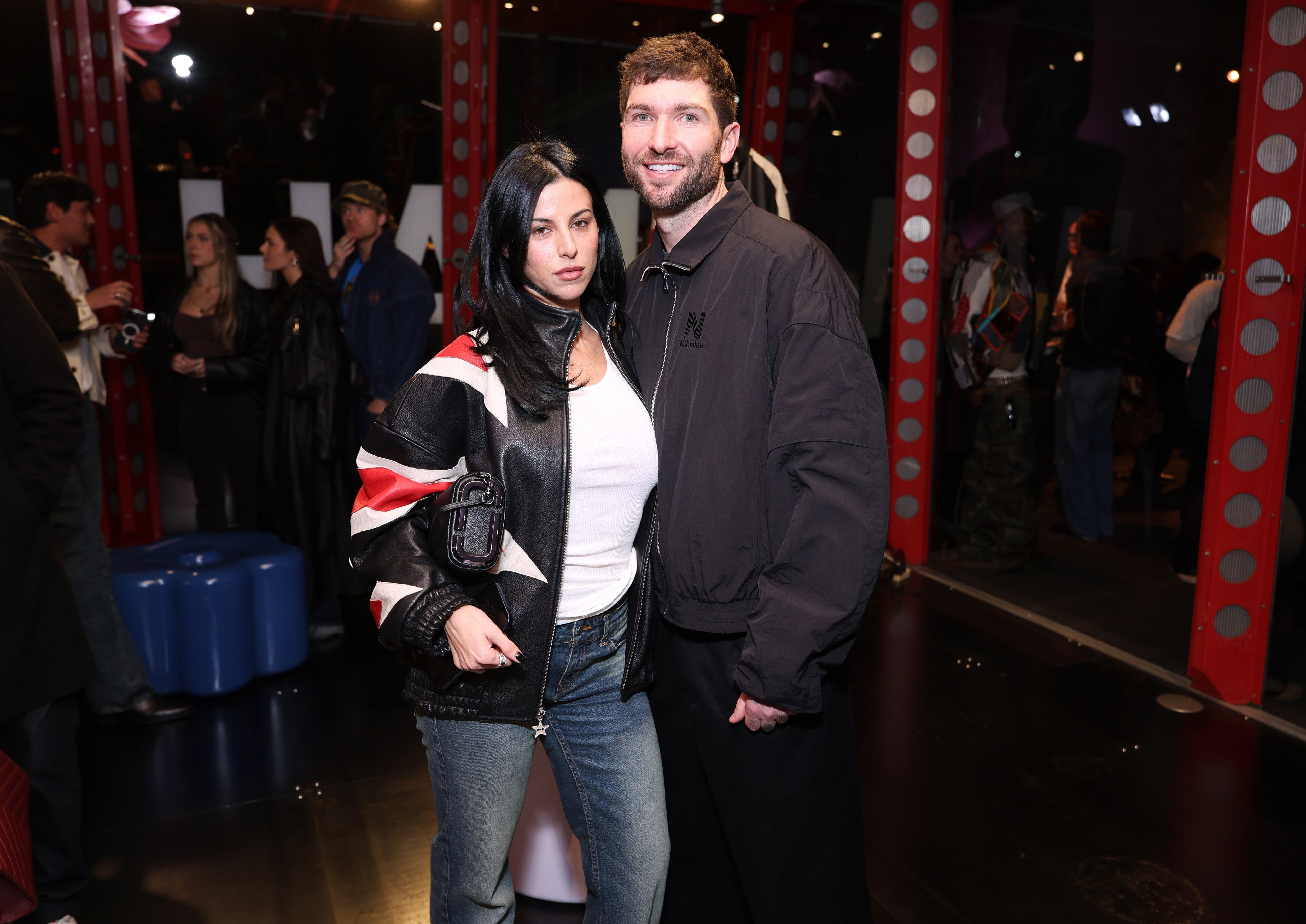 A couple poses at an event; one wears a graphic jacket and jeans, the other wears a dark casual jacket. People socialize in the background