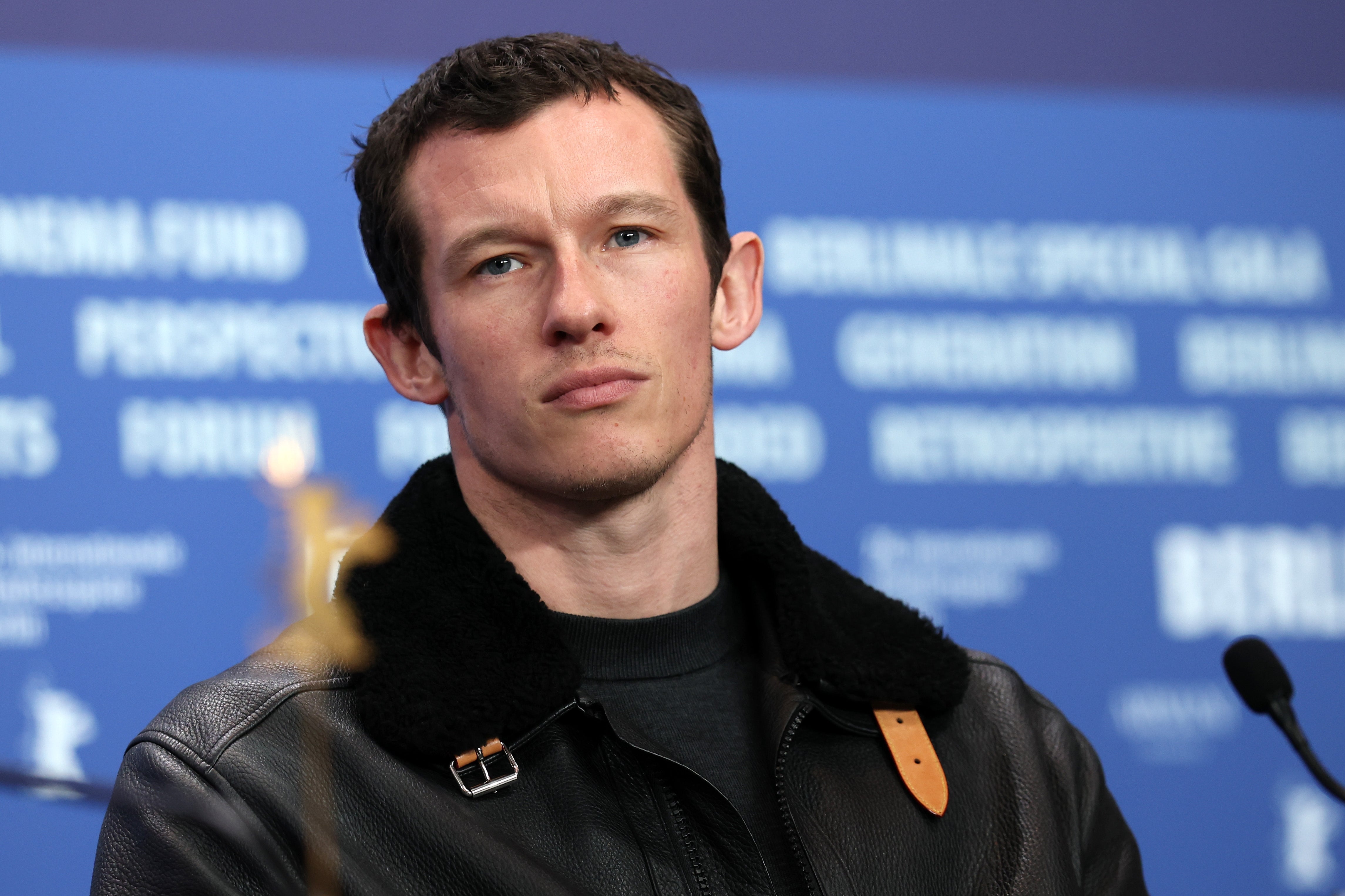 Person at a press event wearing a leather jacket with a shearling collar, looking thoughtful