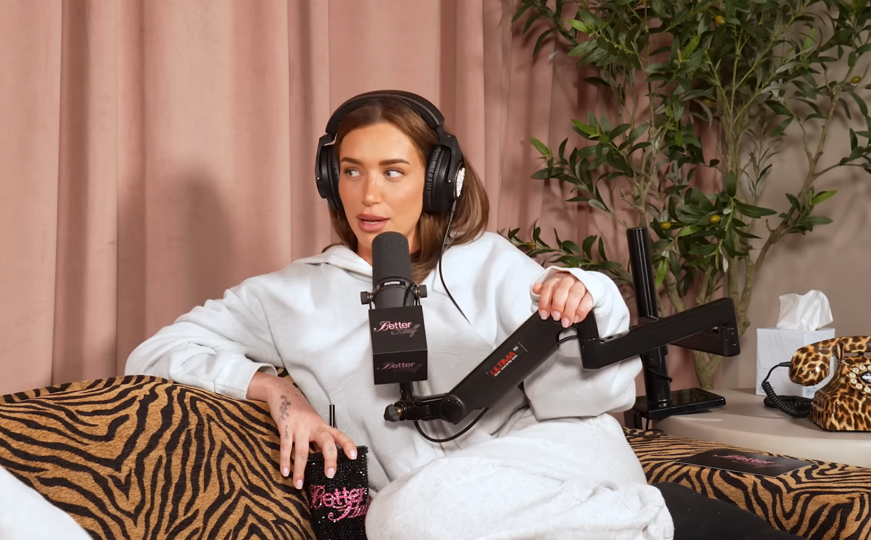 Person in a podcast studio, wearing headphones and a cozy outfit, holding a microphone, seated on a patterned sofa