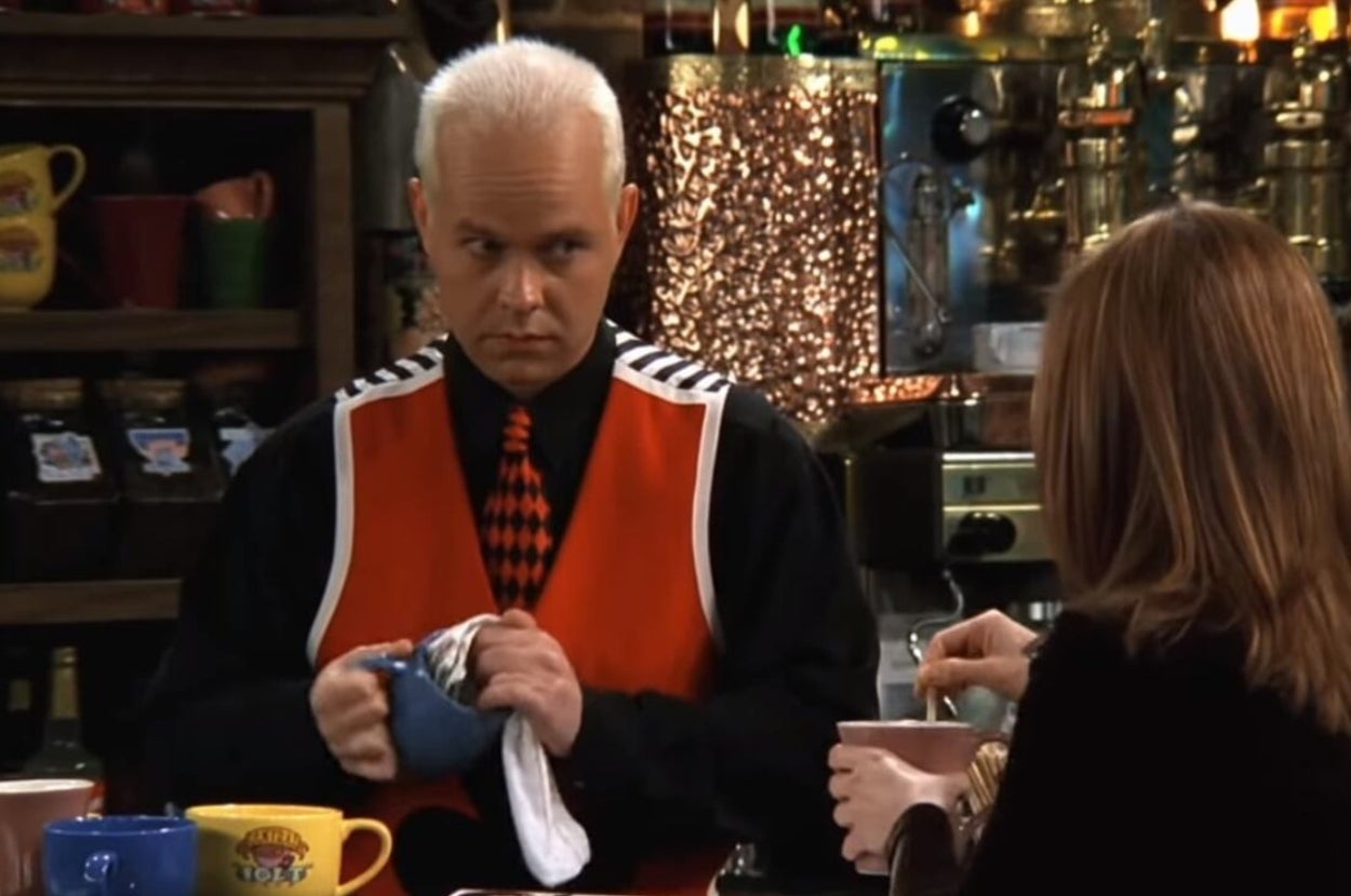 Gunther in "Friends" in a bright vest and checkered tie cleans a mug behind a coffee counter while speaking with Rachel