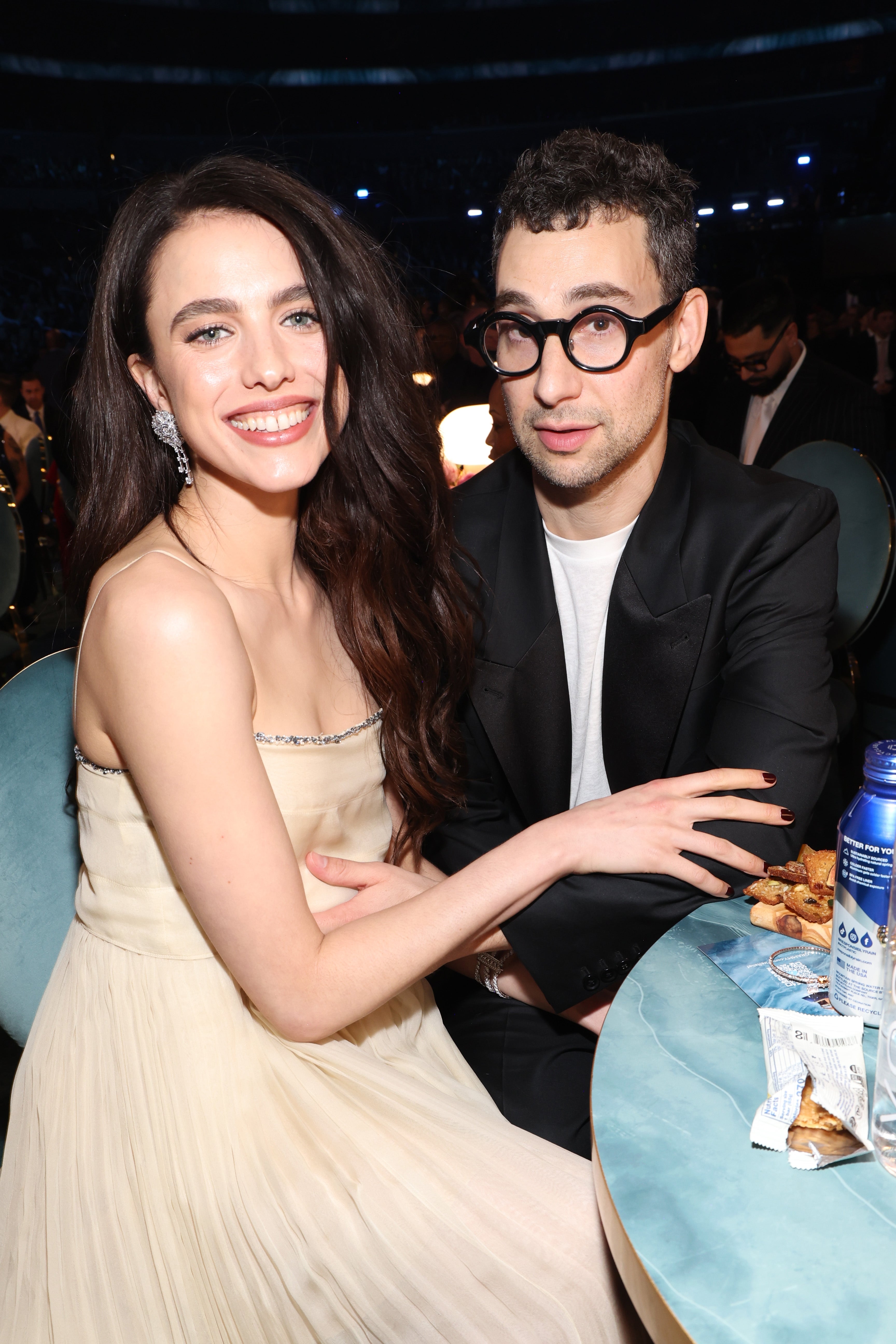 Two people sitting at a table, one in a formal suit and glasses, the other in a sleeveless elegant dress, smiling at an indoor event