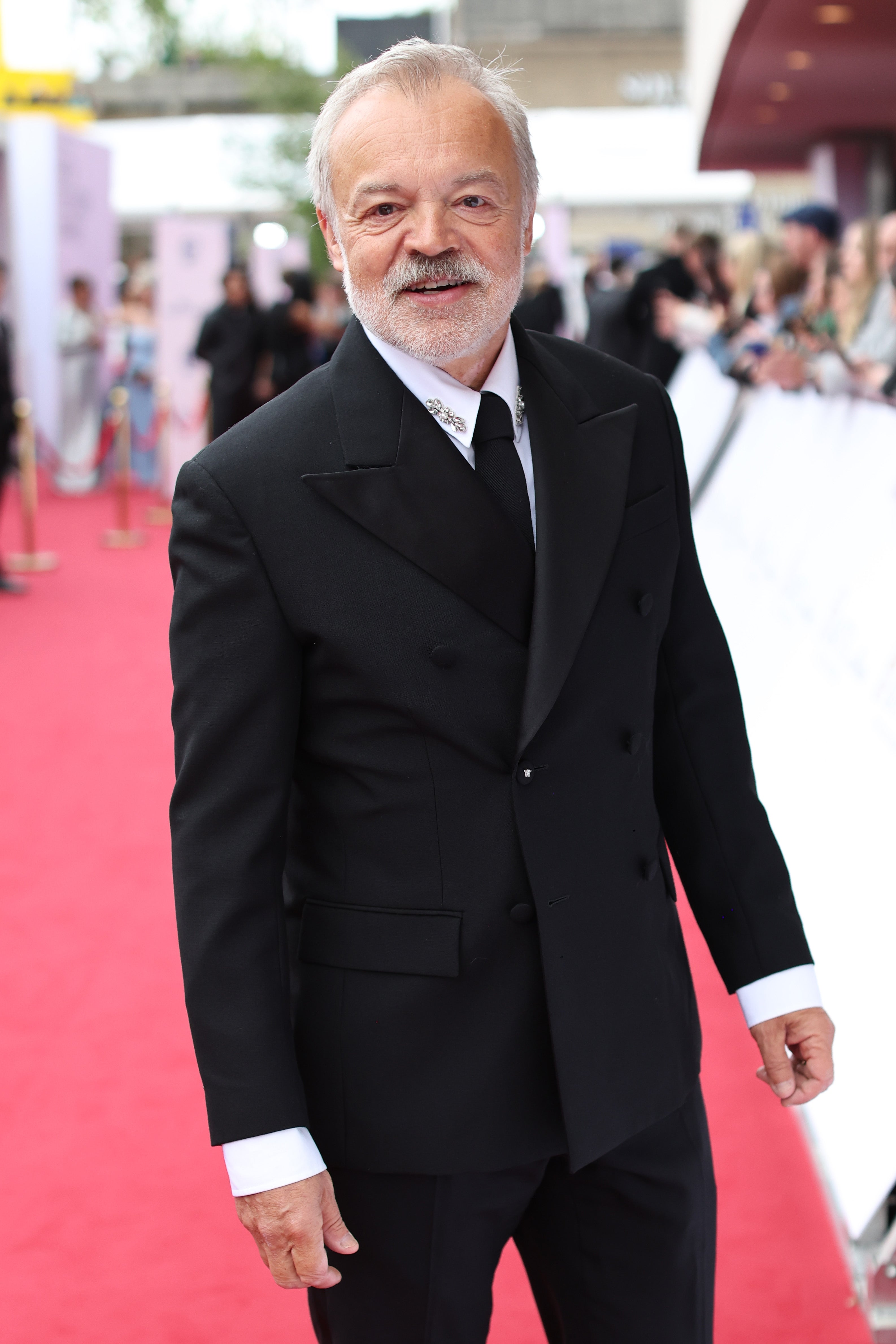 Person in a formal black suit on a red carpet, smiling at the camera. Crowds and photographers are in the background