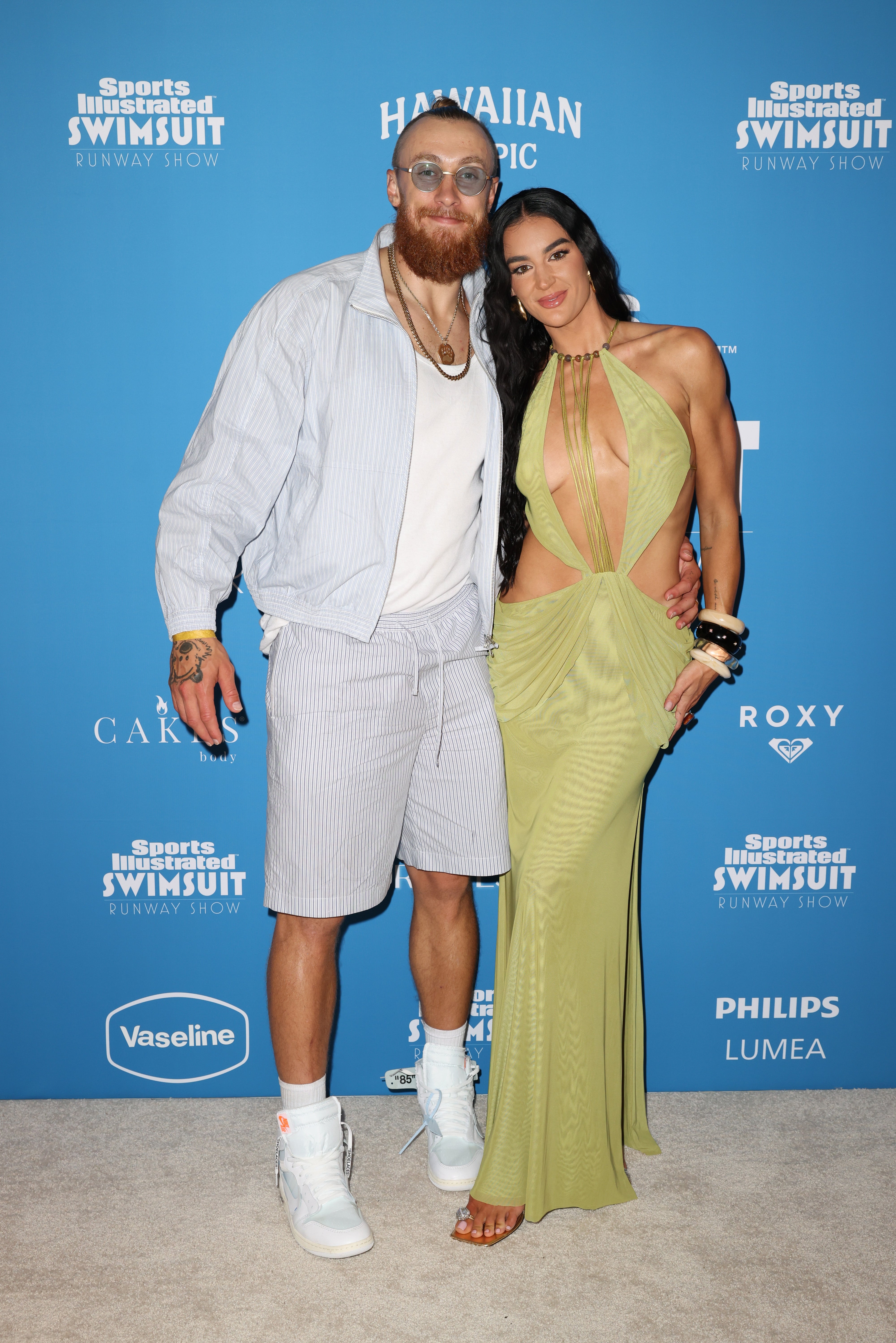 Two individuals on a red carpet; one in a striped suit with sneakers, the other in a flowing, cut-out dress