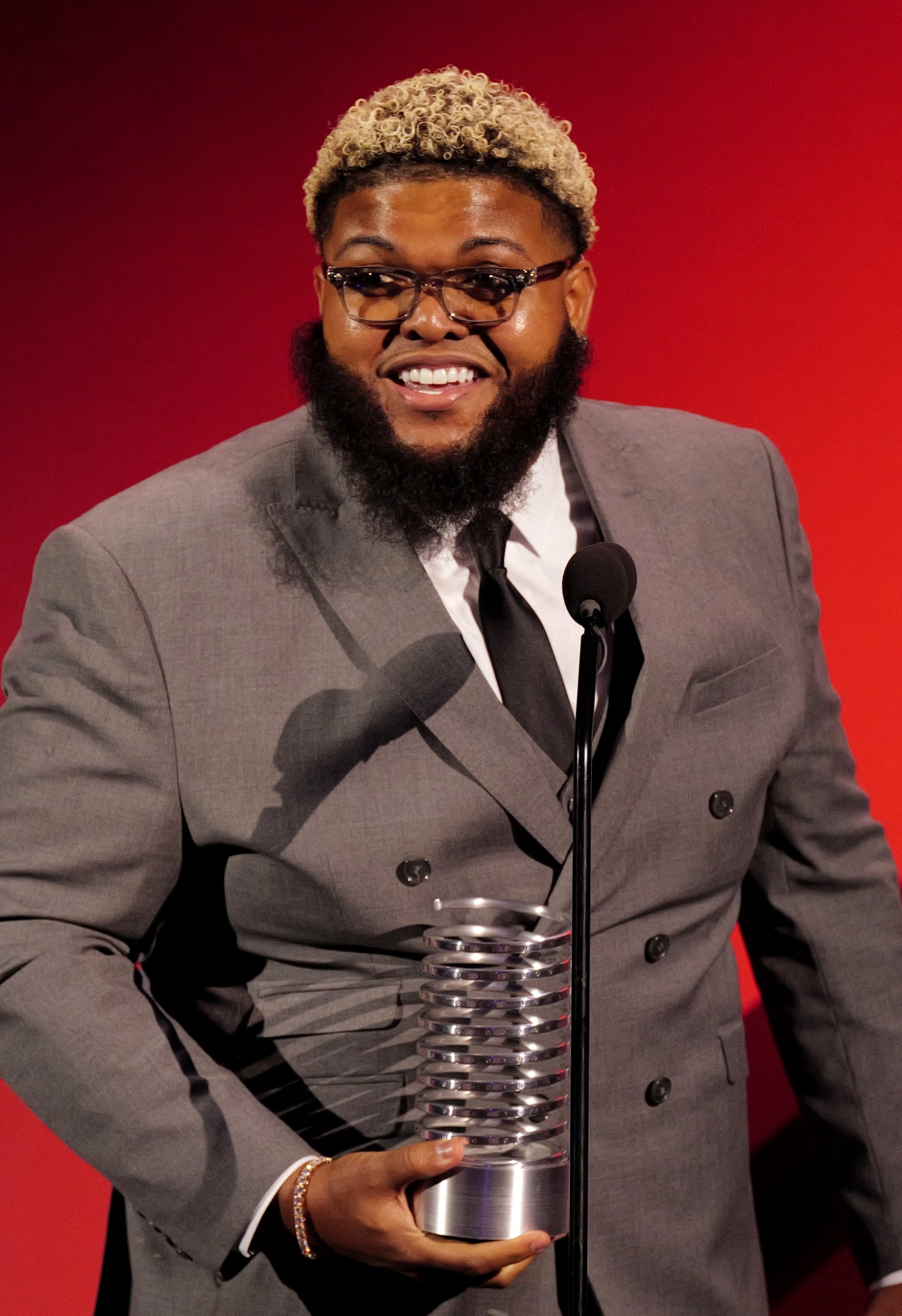 Man in a stylish suit holds an award and speaks at a podium, smiling broadly, likely at an event or ceremony