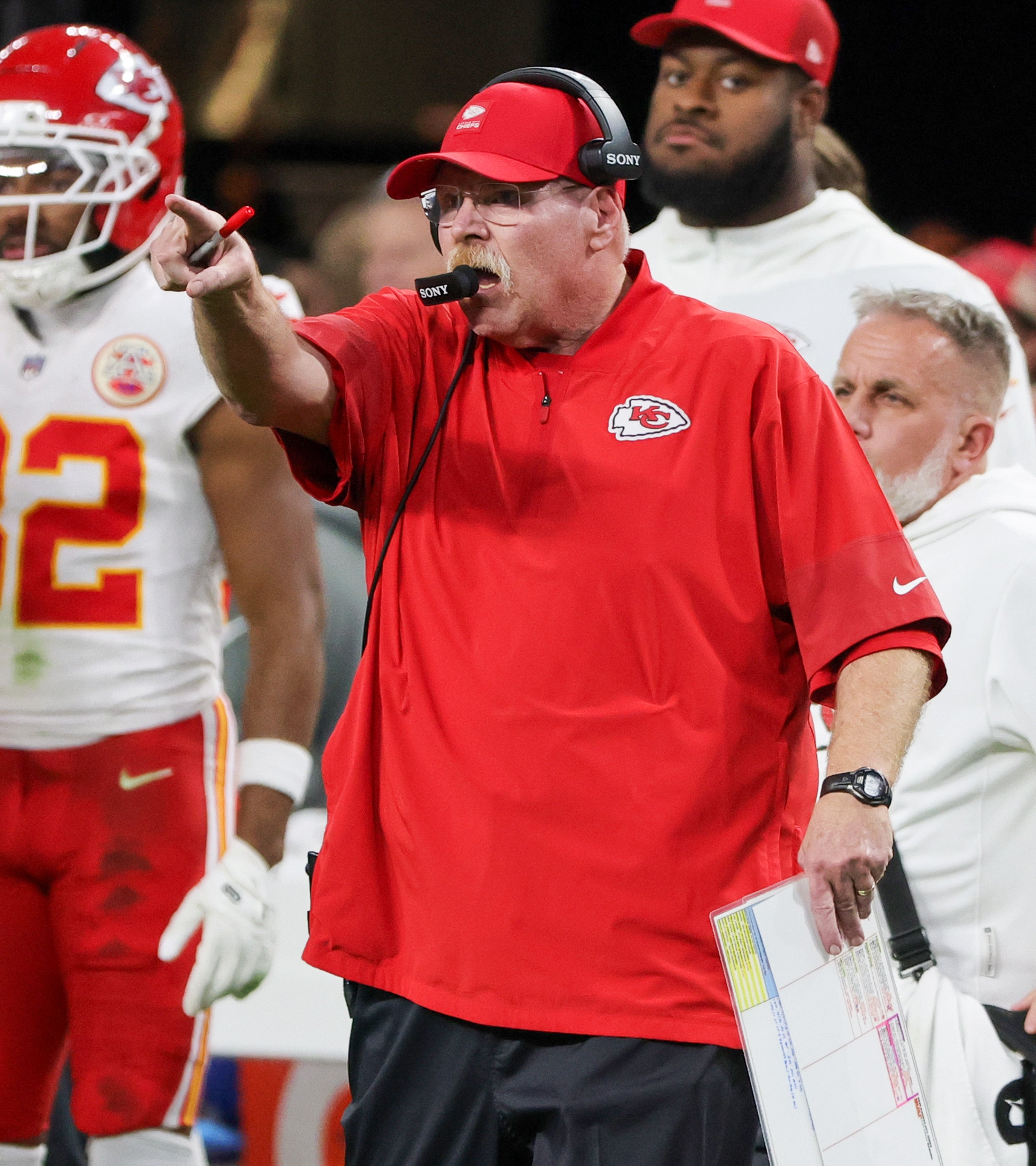 Football coach in a red shirt and cap gestures intensely on the sidelines during a game, surrounded by team members and staff