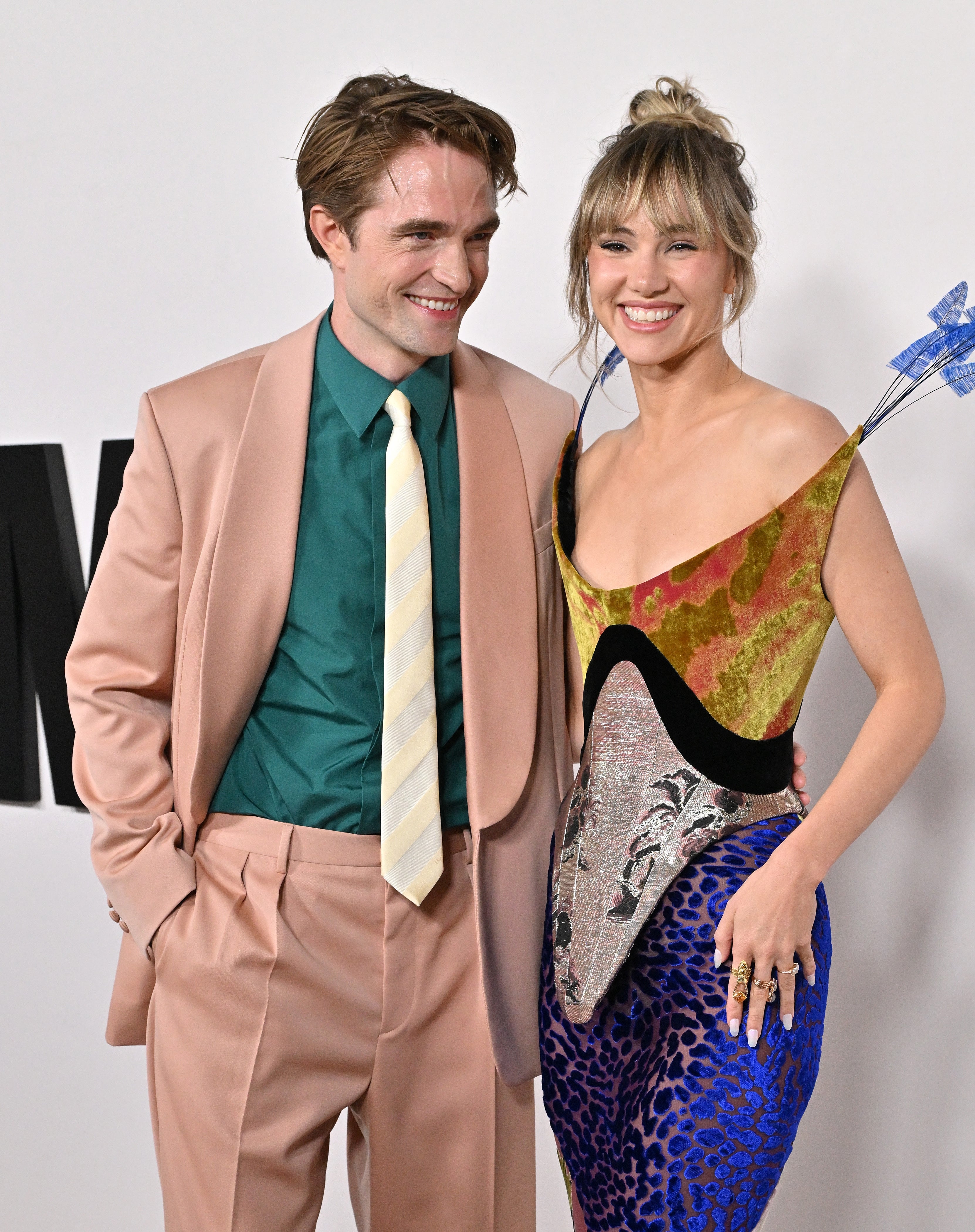 Two people on a red carpet; one in a tan suit with a green shirt, the other in a colorful, patterned dress