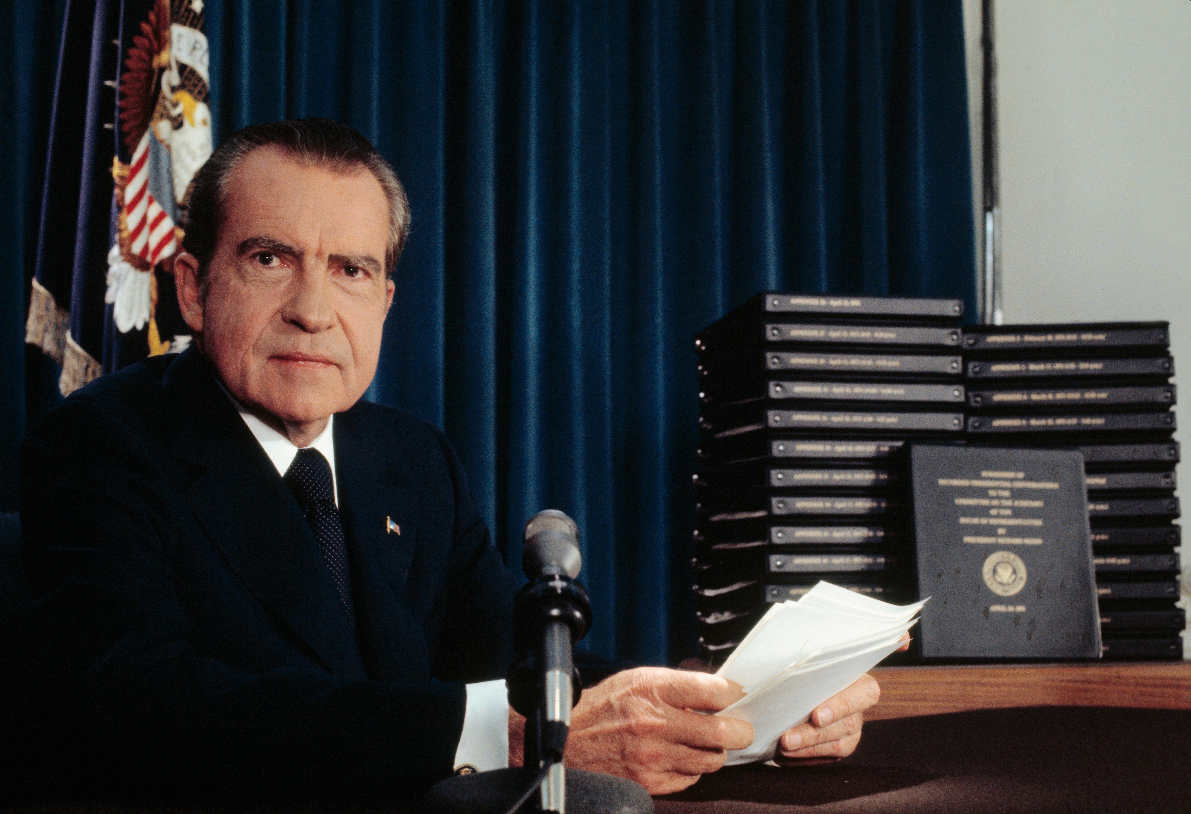A antheral   successful  a suit   sits astatine  a table  with a microphone, holding papers. A ample  stack of binders is to his side