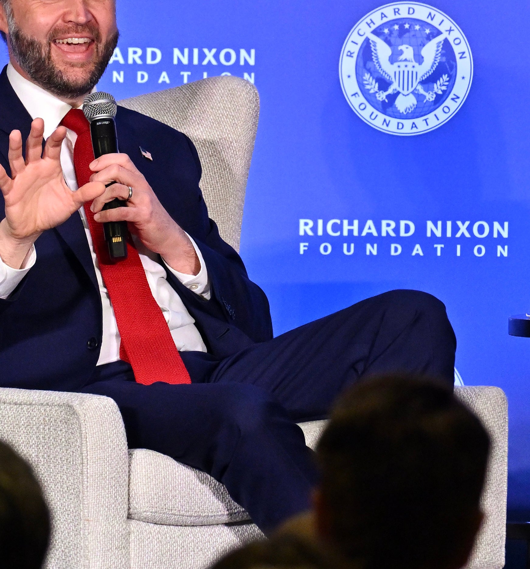Man successful  a suit   seated connected  stage, speaking to an assemblage  astatine  an lawsuit   for the Richard Nixon Foundation. A publication  titled "JD Vance" is connected  the table