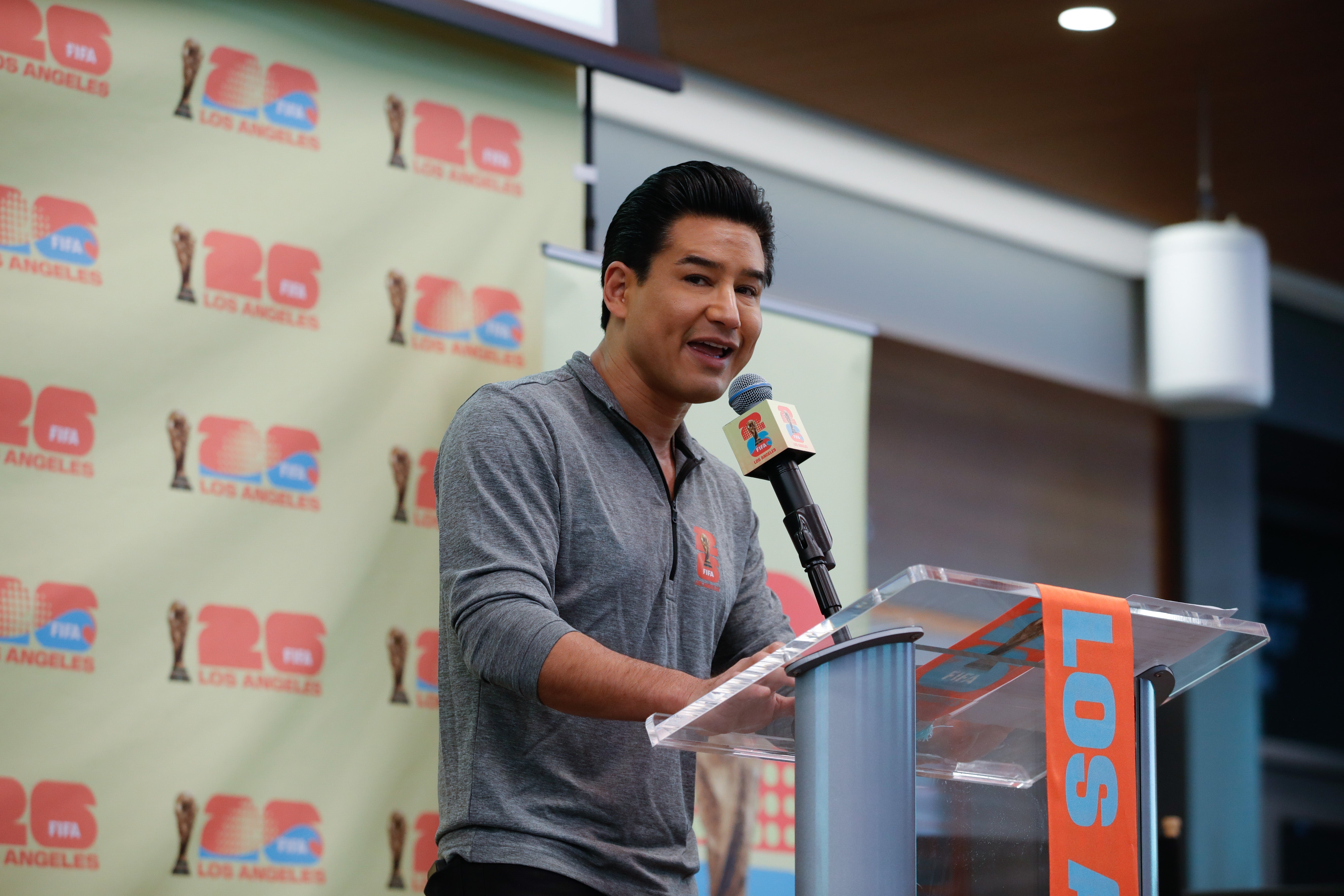 Mario Lopez taler på et podium under en offentlig begivenhet, iført en uformell langermet skjorte, med et bakteppe med logoer
