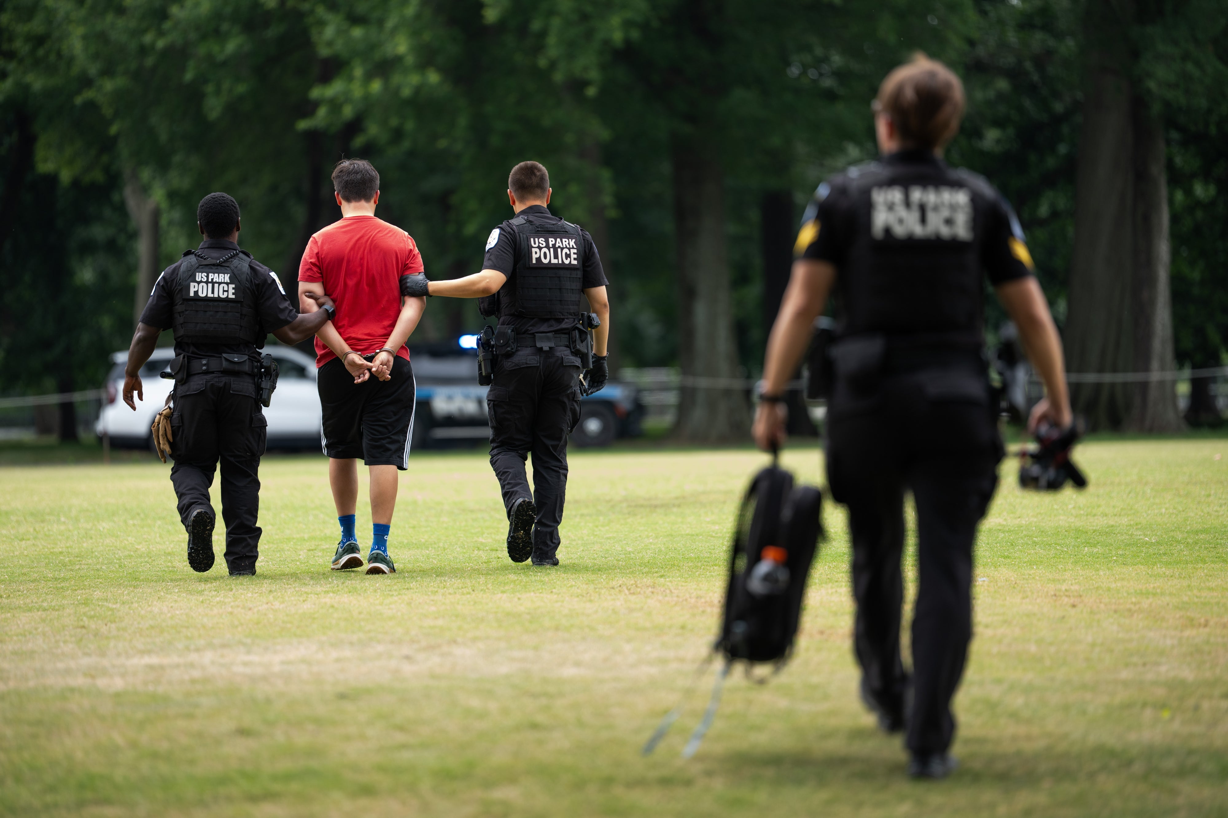 En person i fritidsklær blir eskortert av politifolk gjennom en park. En offiser bærer utstyr og går litt foran