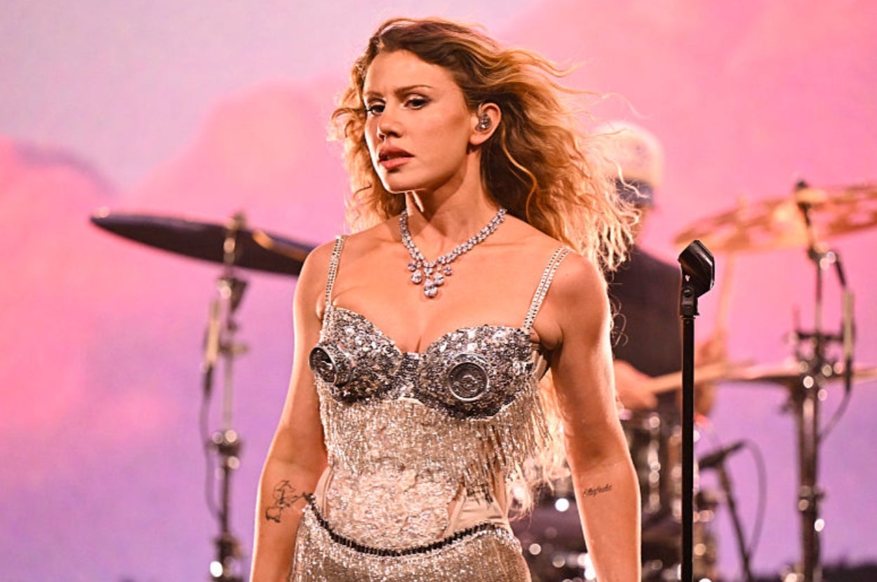 A woman on stage performing, wearing a sparkling, intricate corset dress with fringe. A drummer is visible in the background