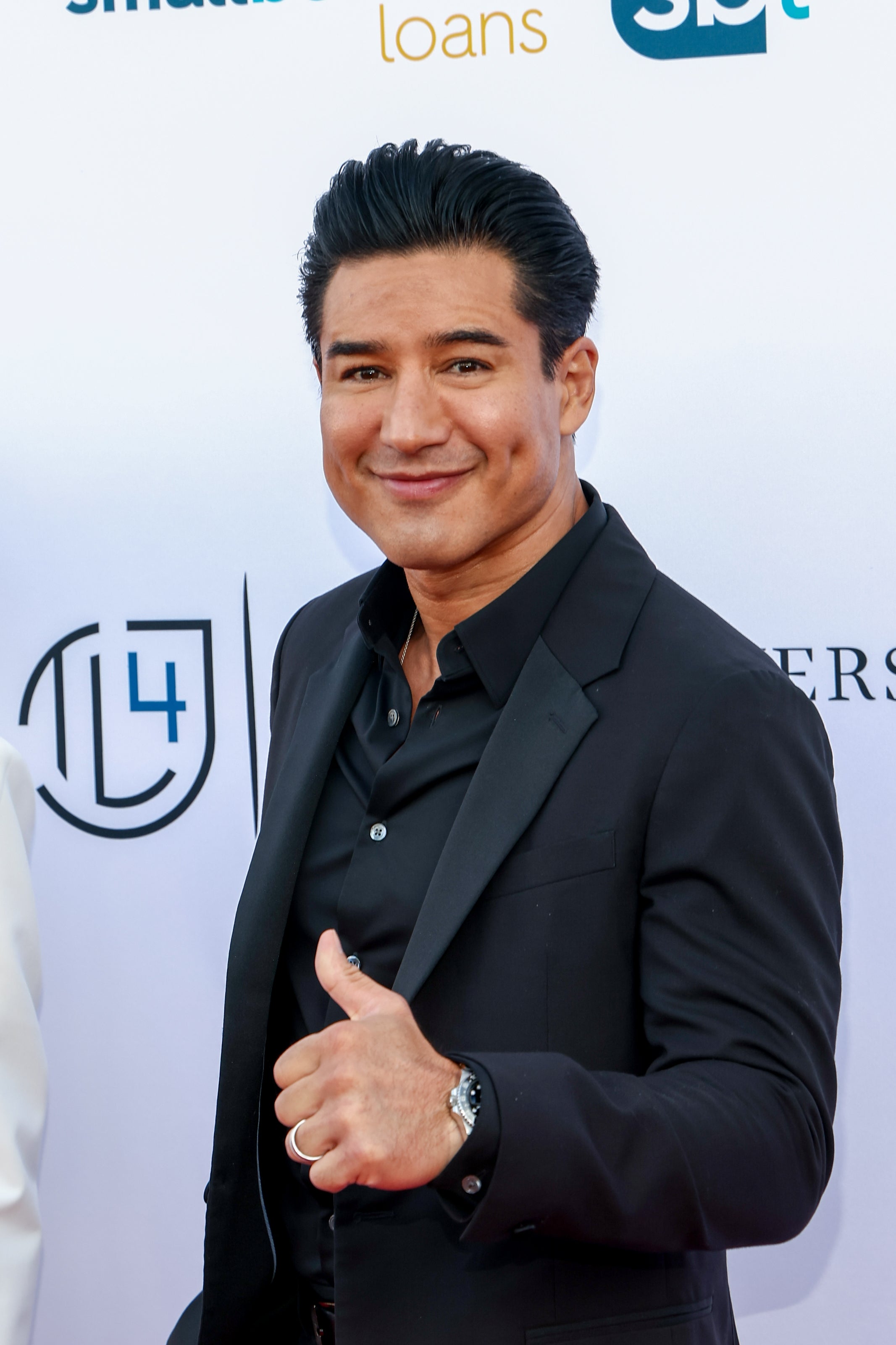 Mario Lopez på rød løper i en elegant dress, smilende med en tommel opp, foran et bakteppe med logoer