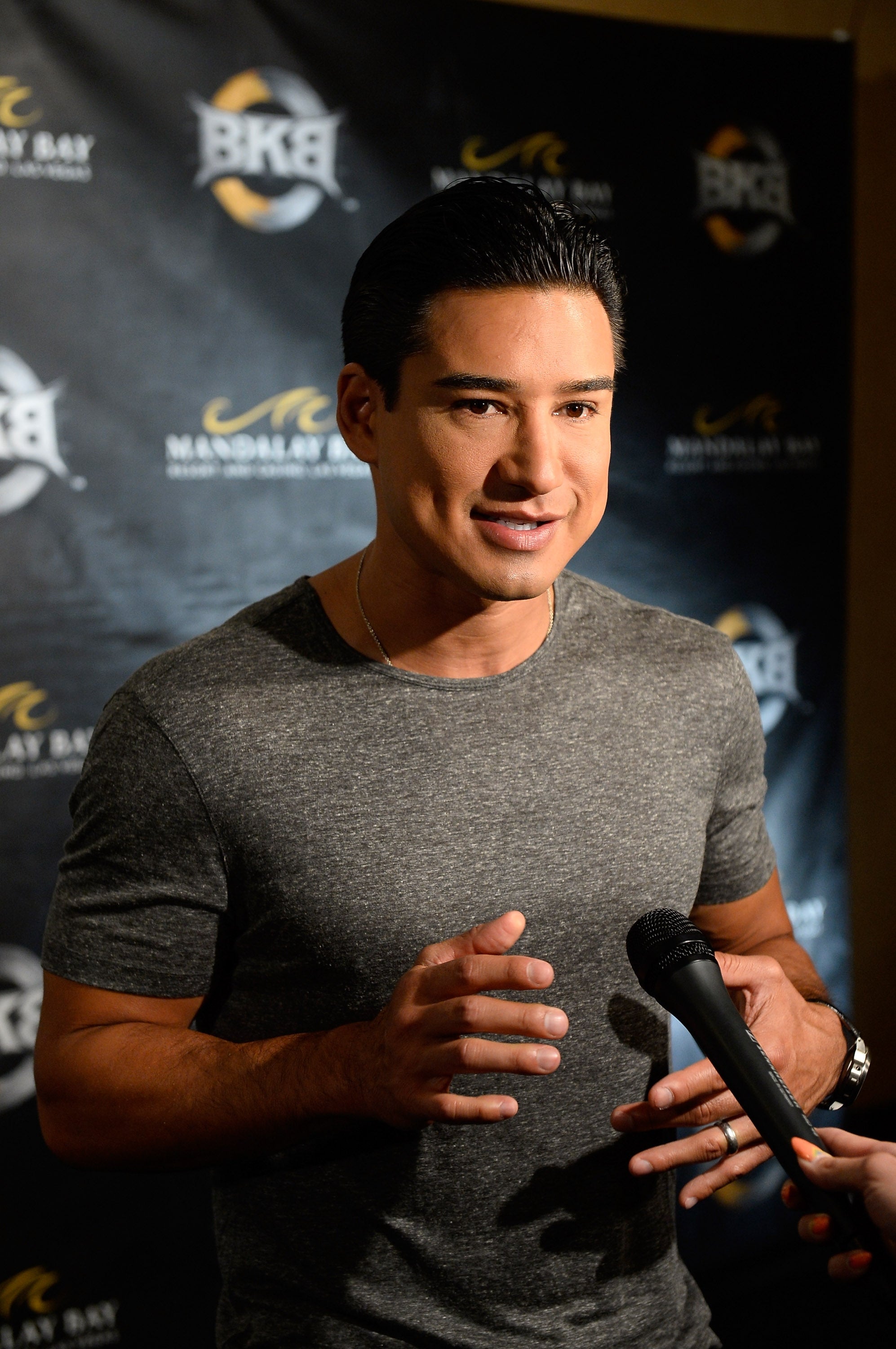 Mario Lopez i en tilpasset t-skjorte snakker på et mediearrangement med en mikrofon i nærheten