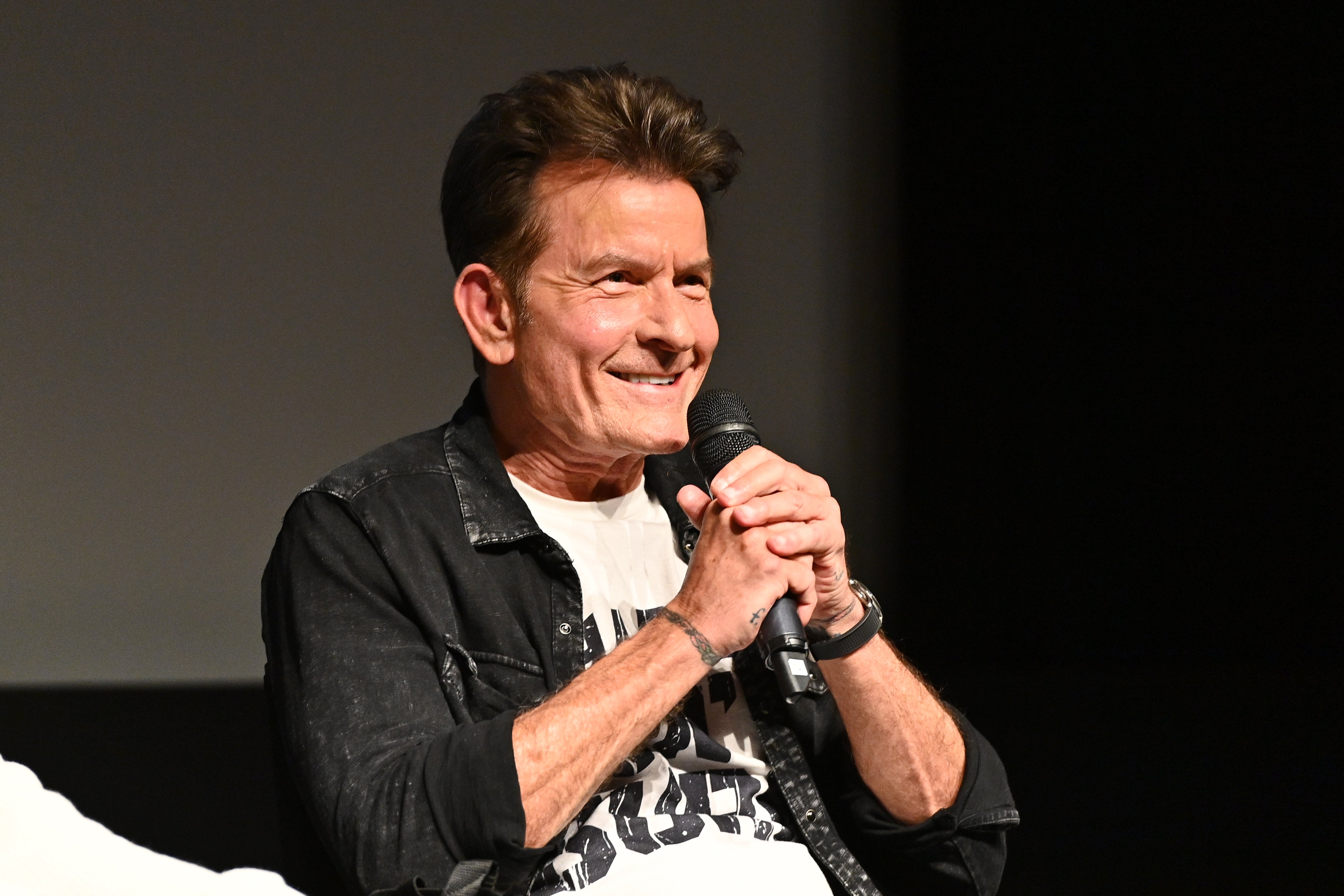 Charlie Sheen smiling and holding a microphone, wearing a casual jacket over a graphic t-shirt, seated on stage during a panel discussion