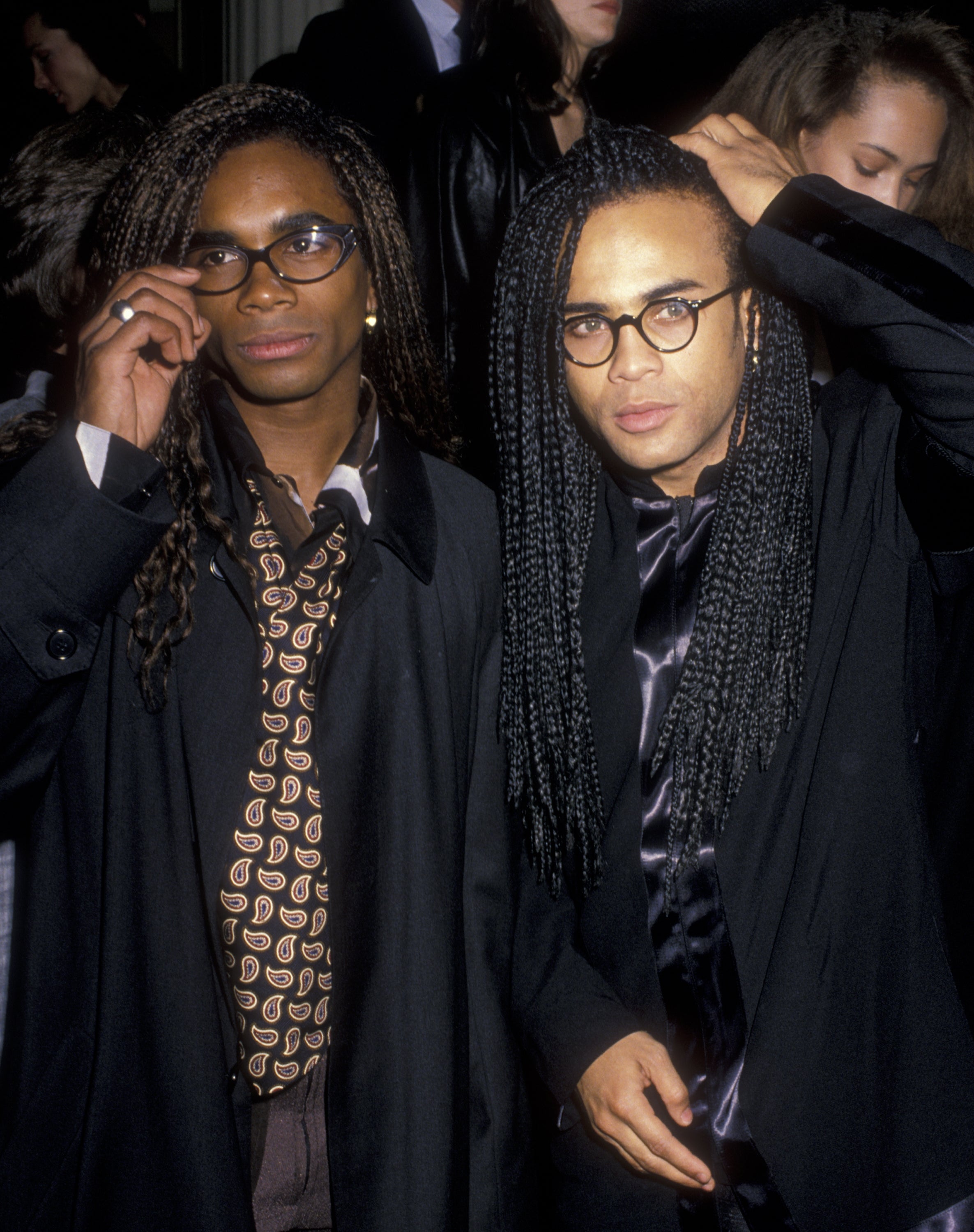 Milli Vanilla with long braided hair wearing glasses and stylish coats at an event
