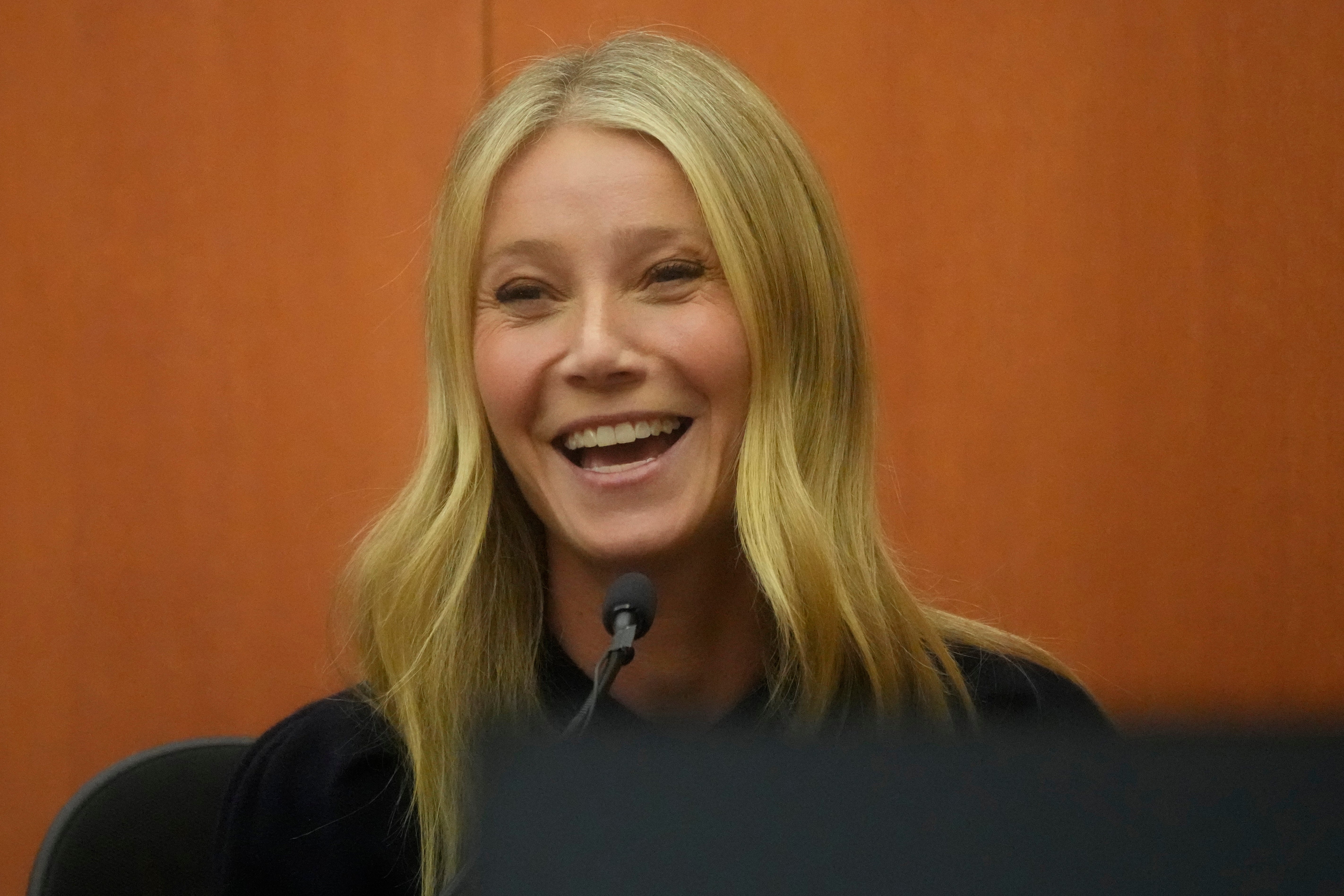 Gwyneth Paltrow with long hair smiling while speaking into a microphone at an indoor event