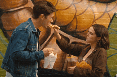 Two people enjoying takeaway food in front of a mural. One person feeds the other with a fried item, both holding takeout containers