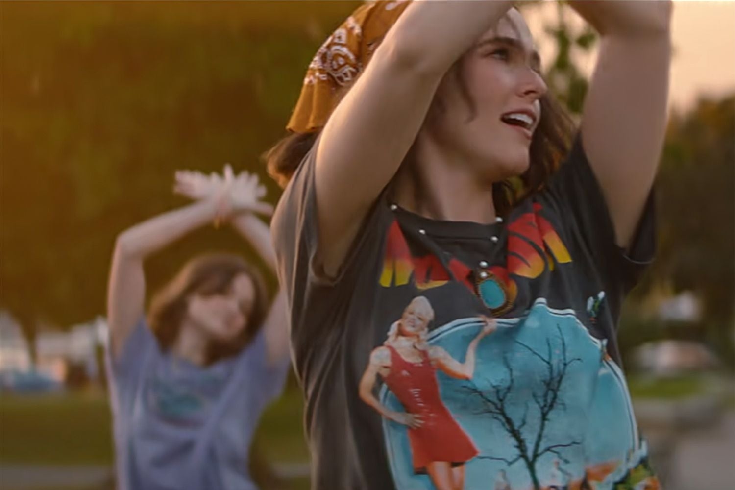 Two people dancing outdoors, smiling. The one in front wears a graphic tee and bandana, the other in the background is slightly blurred