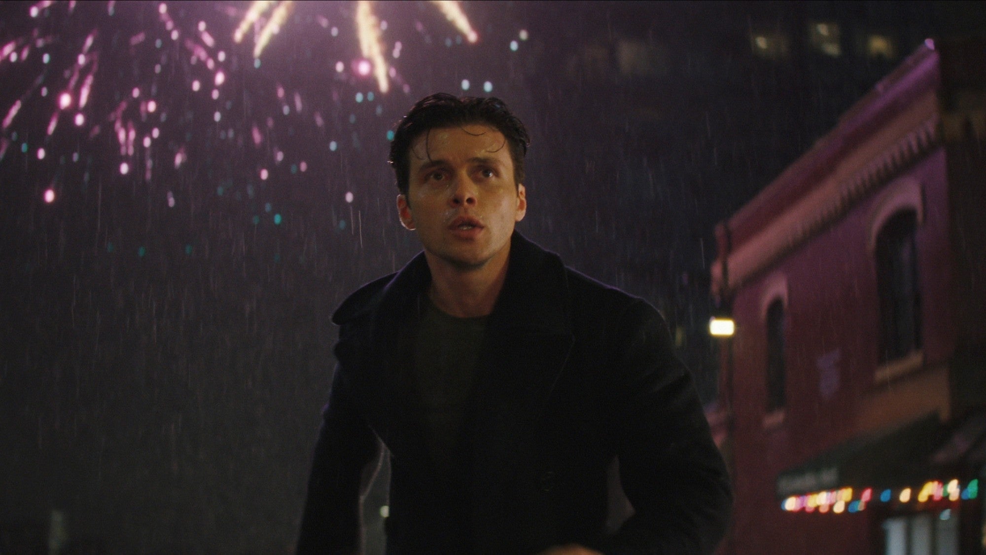 A person looks up at fireworks in the night sky, standing in front of a lit building, appearing surprised or thoughtful