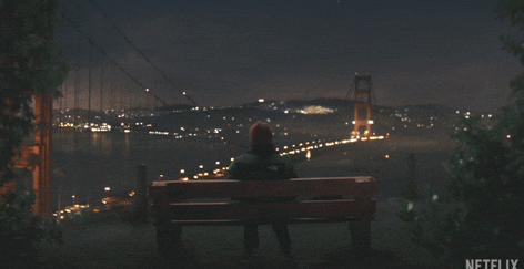 Person sitting on a bench overlooking a lit-up suspension bridge at night, surrounded by a calm, scenic setting