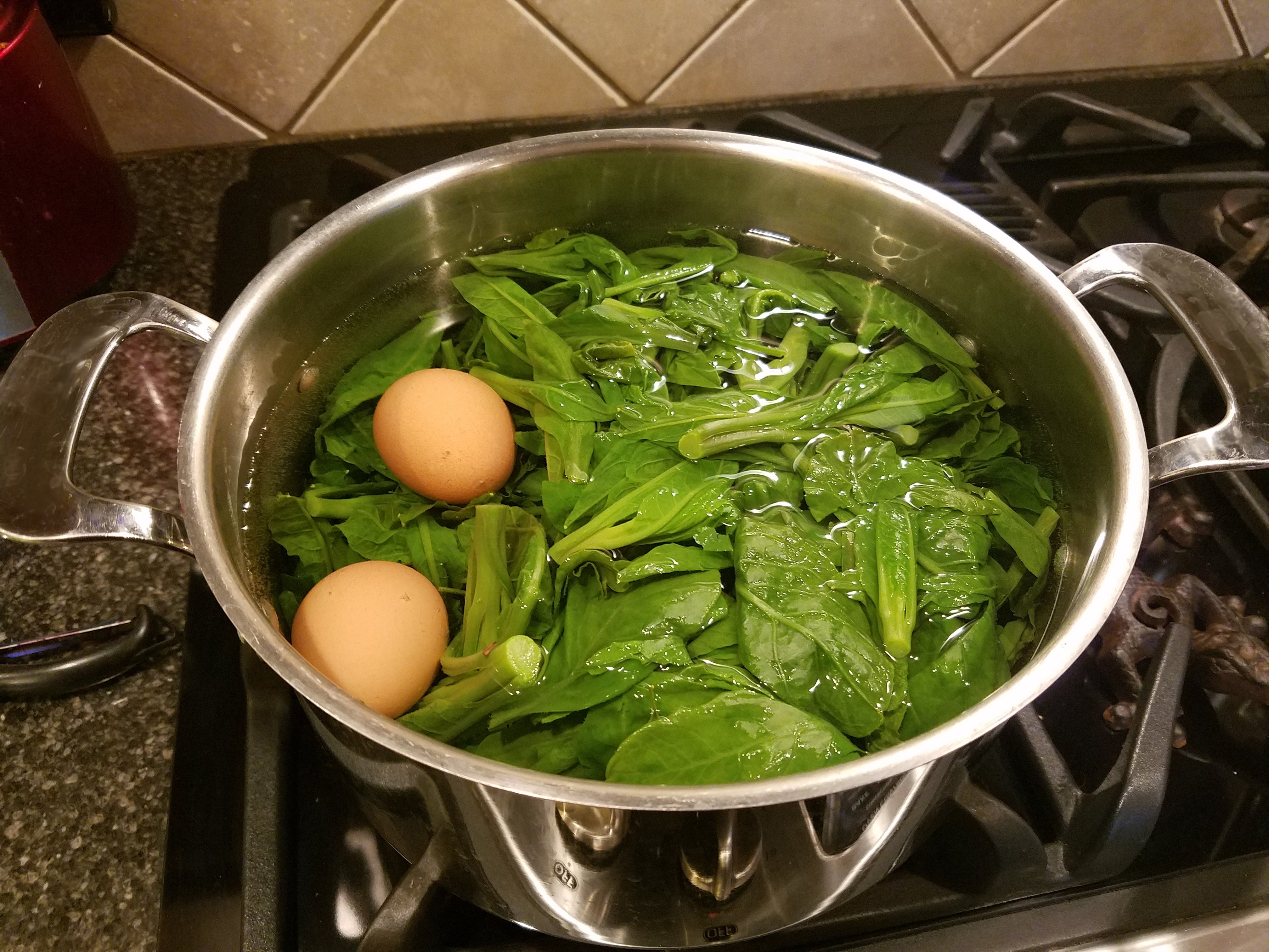Pot of spinach and 2 eggs being boiled connected a stovetop