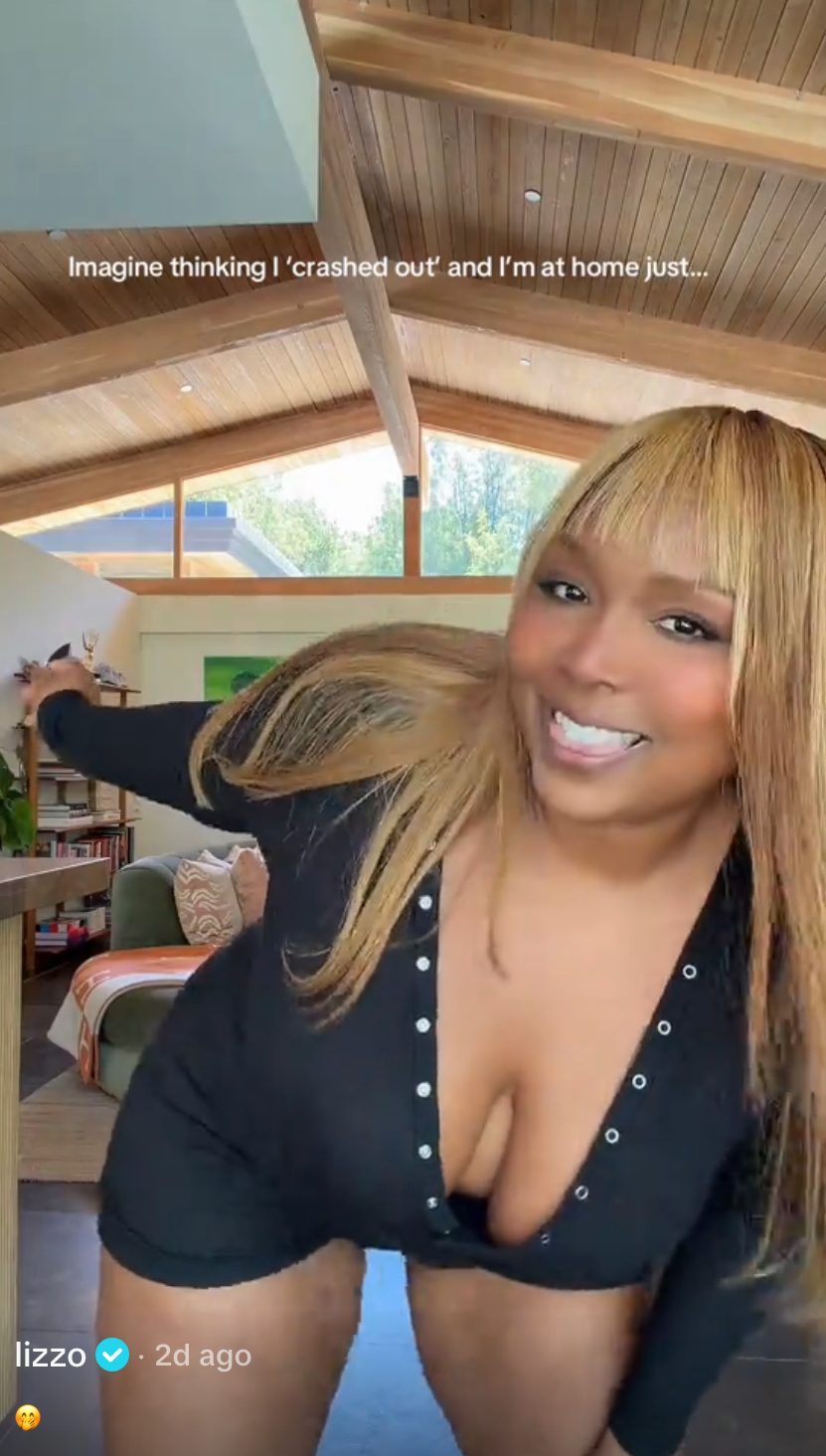 Lizzo smiling and posing at home in a V-neck outfit with button details, text above reads: "Imagine thinking I 'crashed out' and I'm at home just..."
