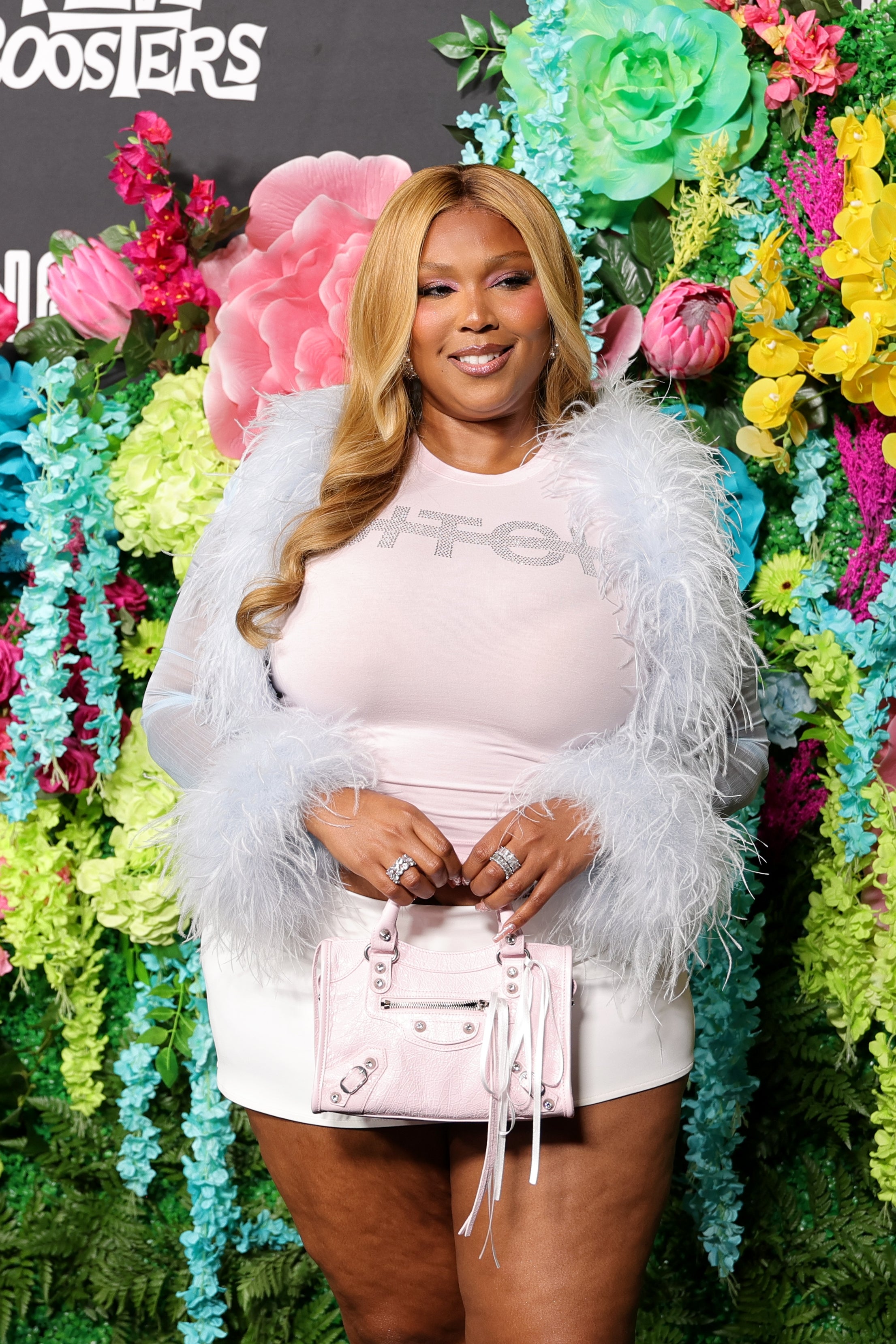 Lizzo in feathered top and mini skirt smiles against a floral backdrop, holding a small handbag